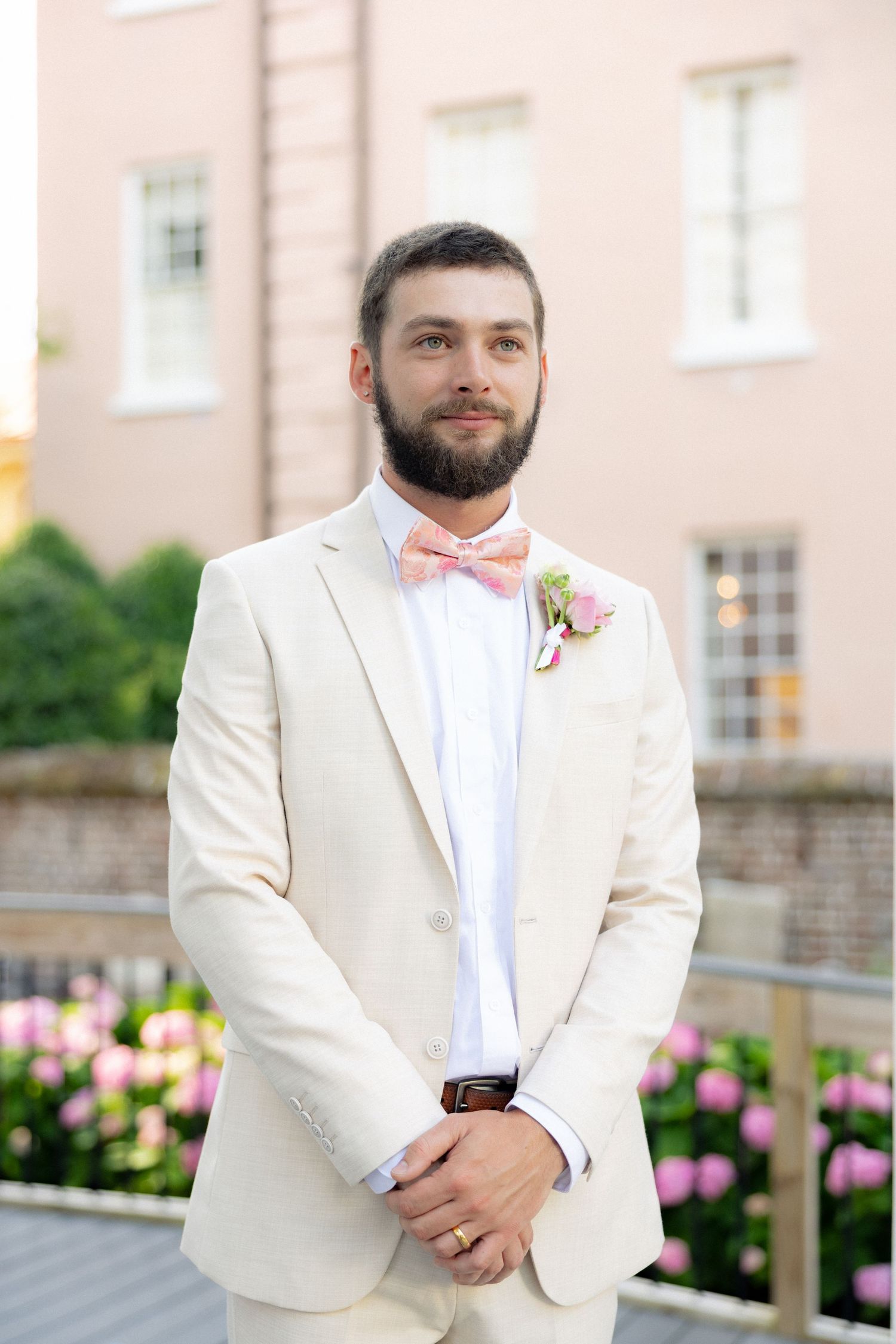 Bearded groom in cream suit with pink bow tie and floral boutonniere standing outdoors.