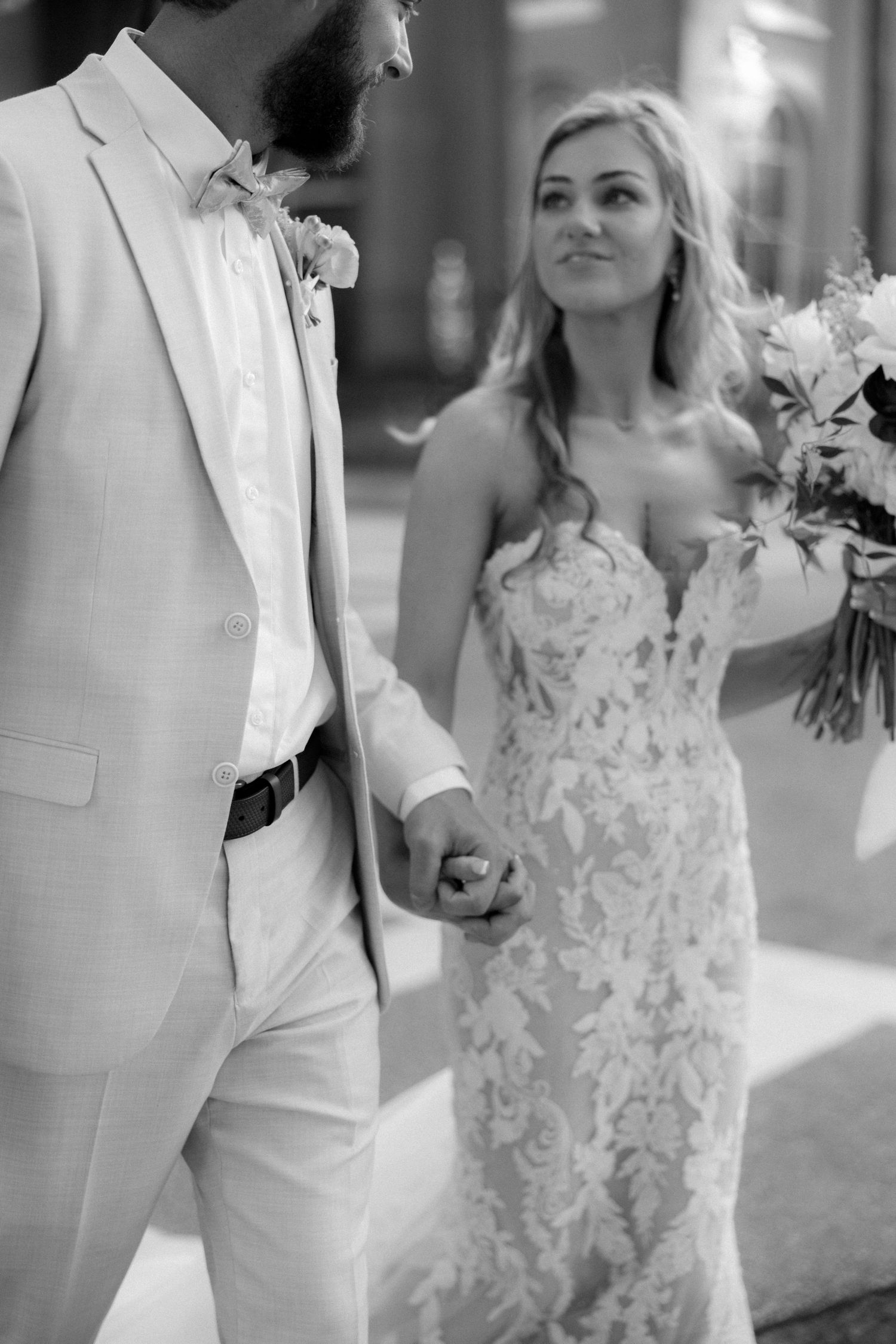 Black and white photo of bride and groom holding hands, bride in lace gown holding bouquet.