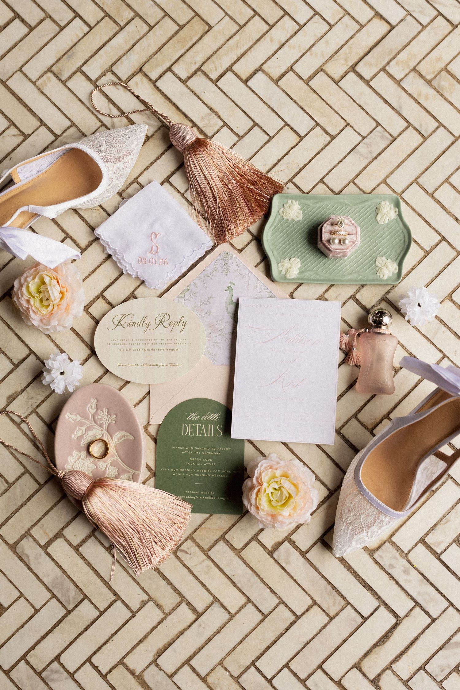 Elegant wedding flat lay with blush invitations, lace heels, pink tassels, floral accents, and sage green details on herringbone surface.