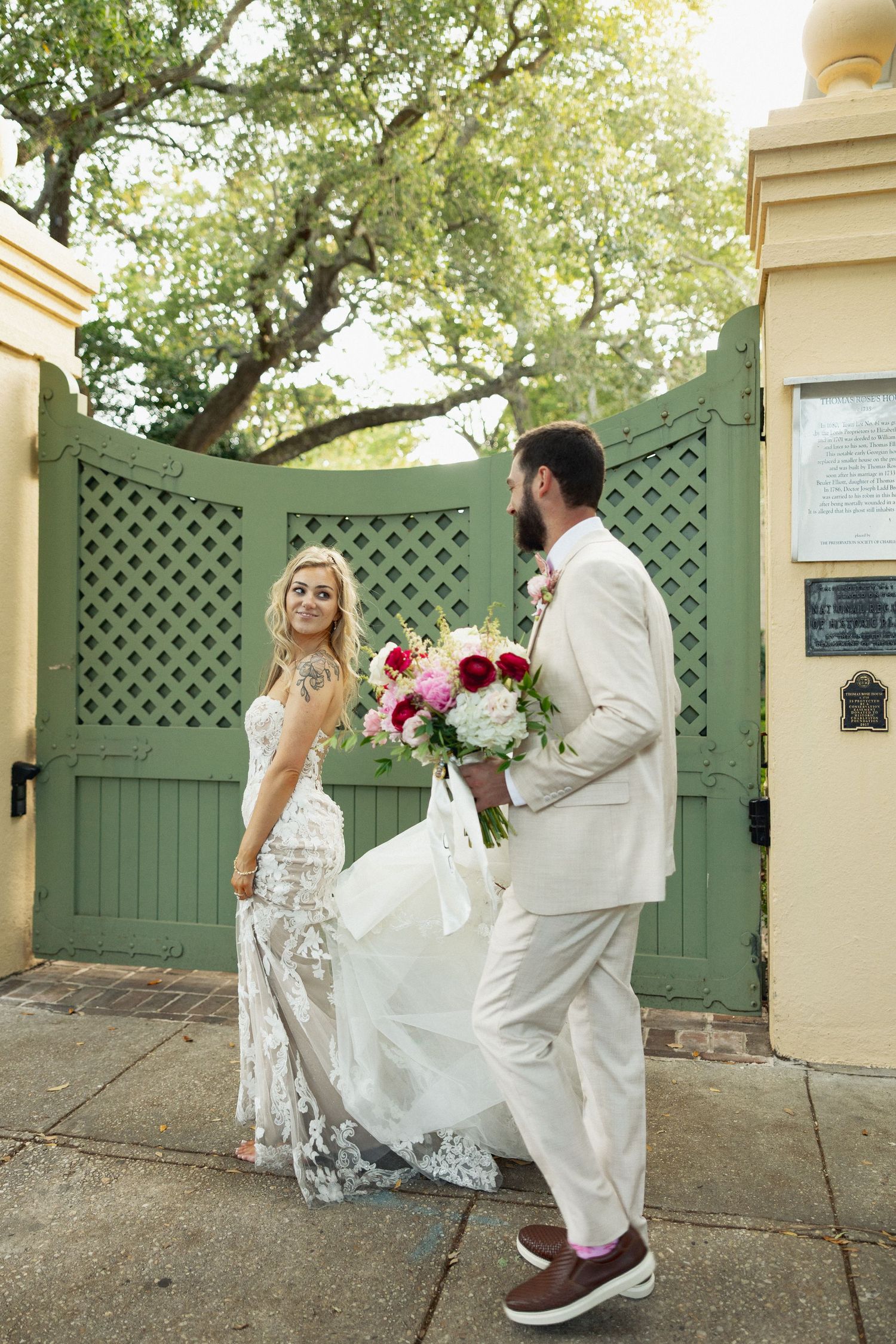 Bride and groom walking through green lattice gate, bride in lace gown holding pink bouquet, groom in white suit.