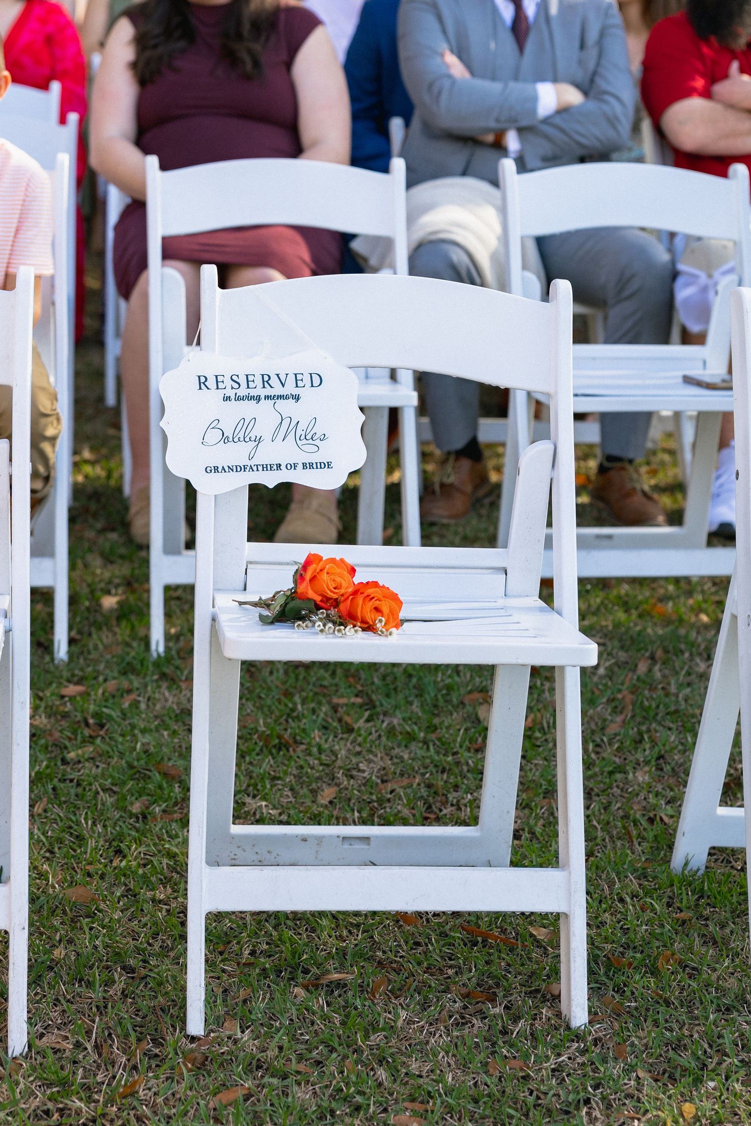 White reserved chair with orange roses honoring Billy Miles, grandfather of the bride, at a wedding ceremony.