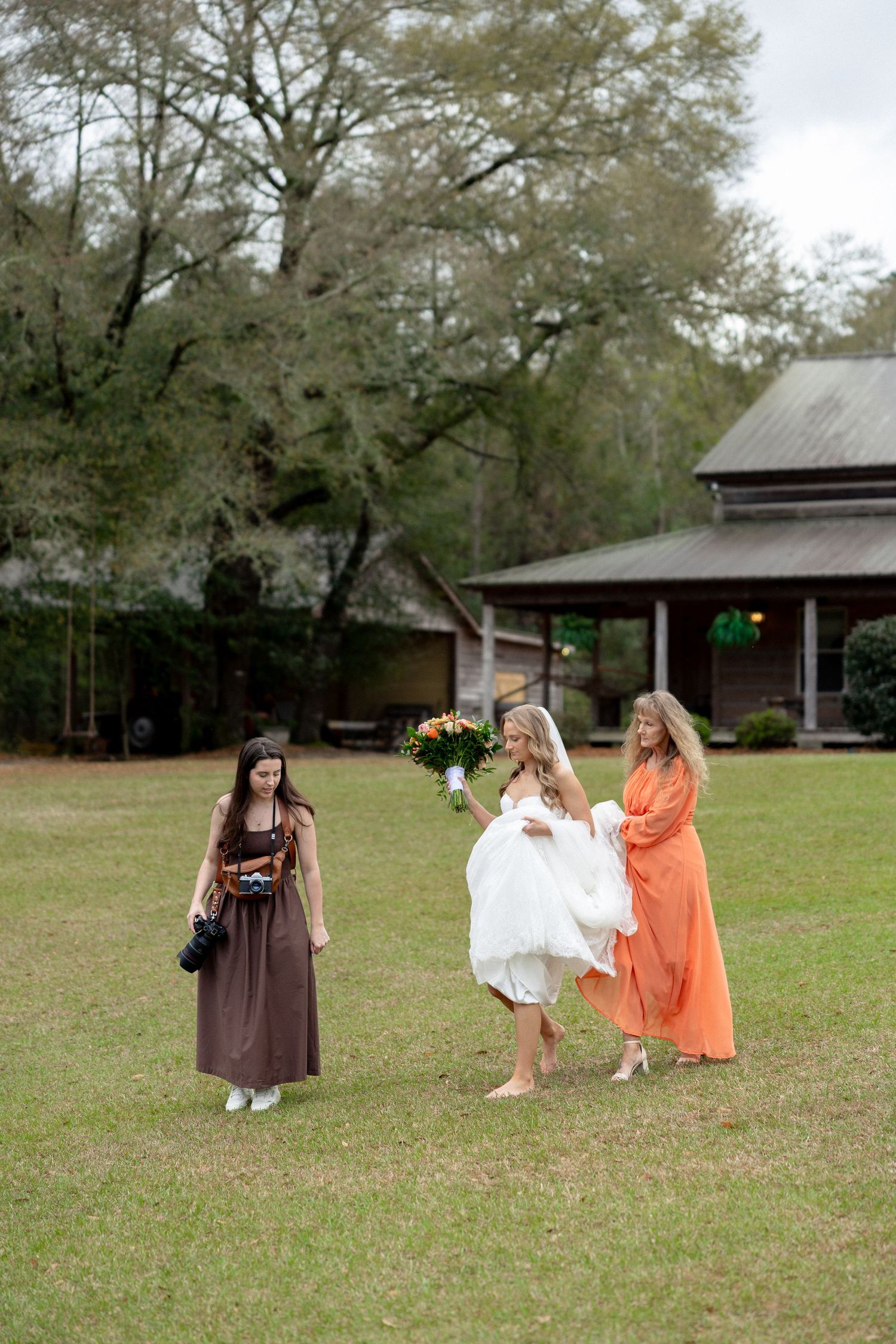 Bride in white wedding gown holding bouquet walks on lawn while photographer captures the moment.