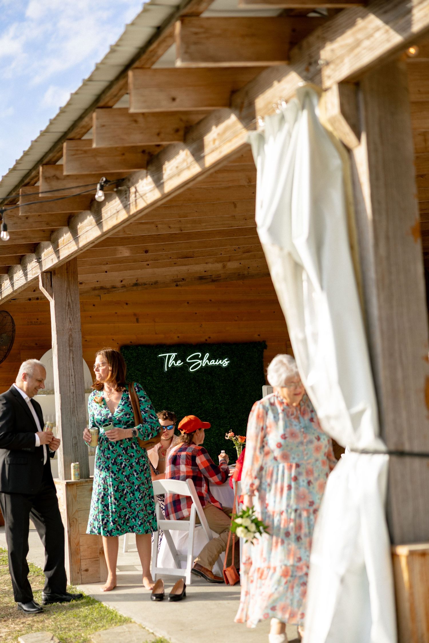 Wedding guests mingling under rustic wooden pavilion with The Shaws green neon sign backdrop.