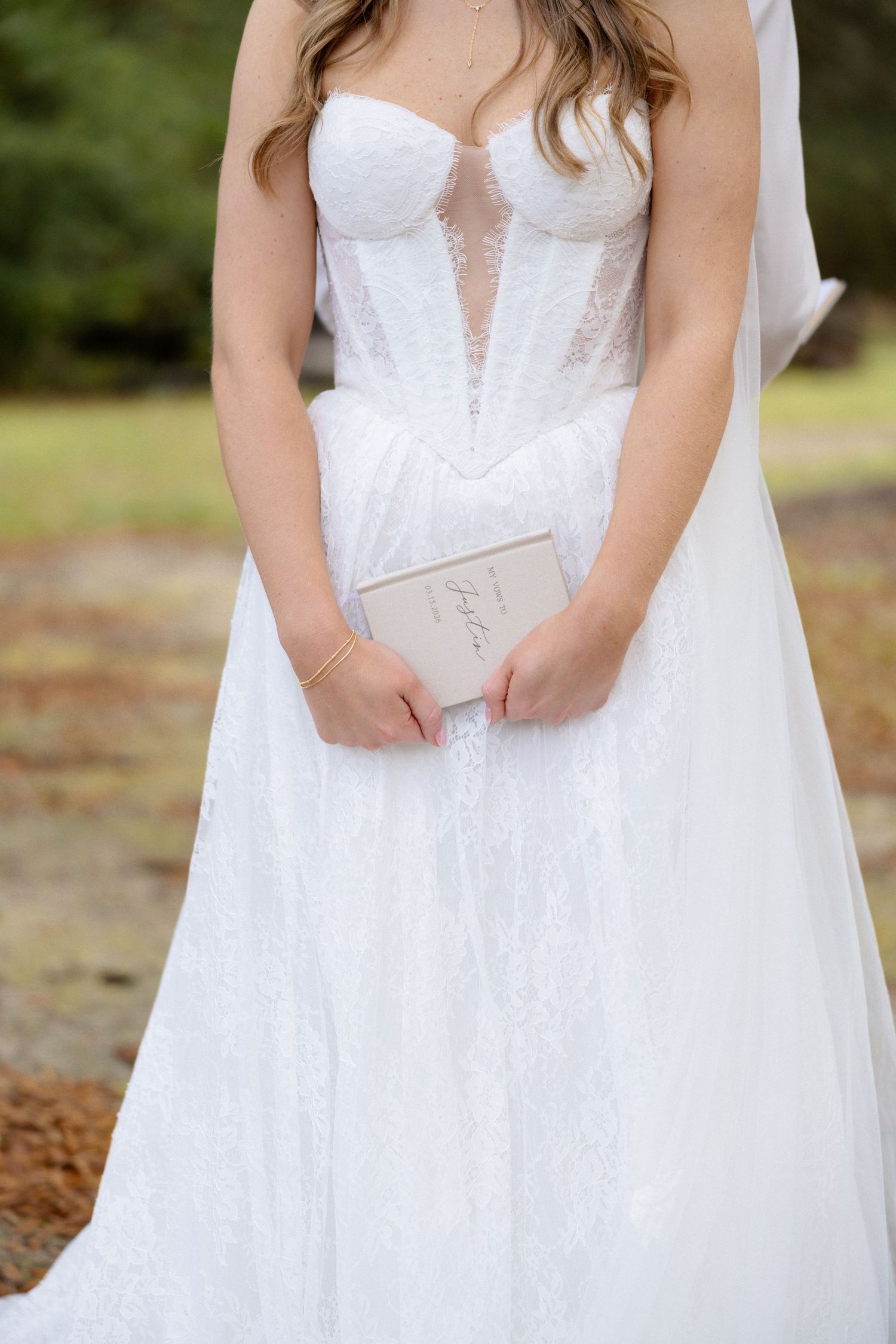 Close-up of bride in strapless lace corset wedding gown holding beige vow book outdoors.
