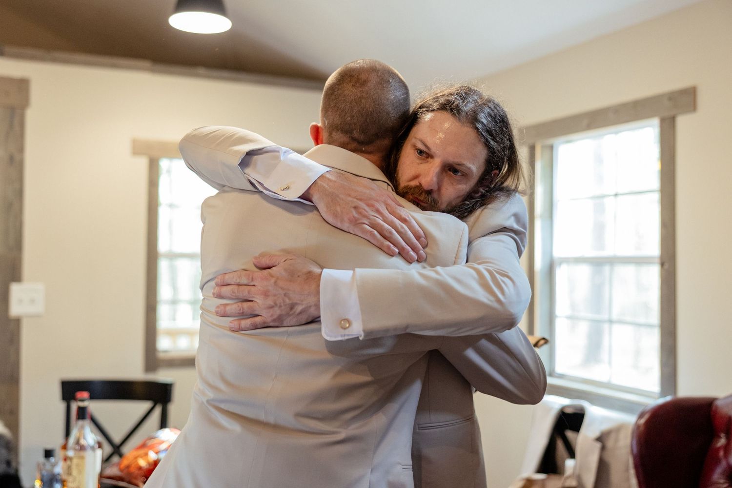 Two groomsmen in beige suits share a warm embrace during wedding day preparations indoors.