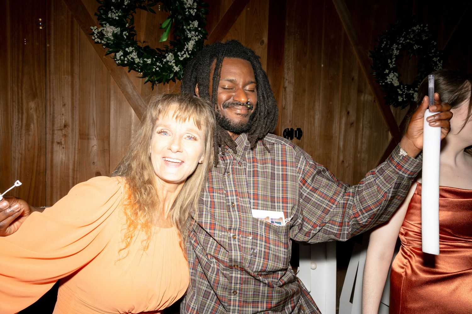 Happy wedding guests, a woman in orange and a man in plaid, pose together at barn reception.