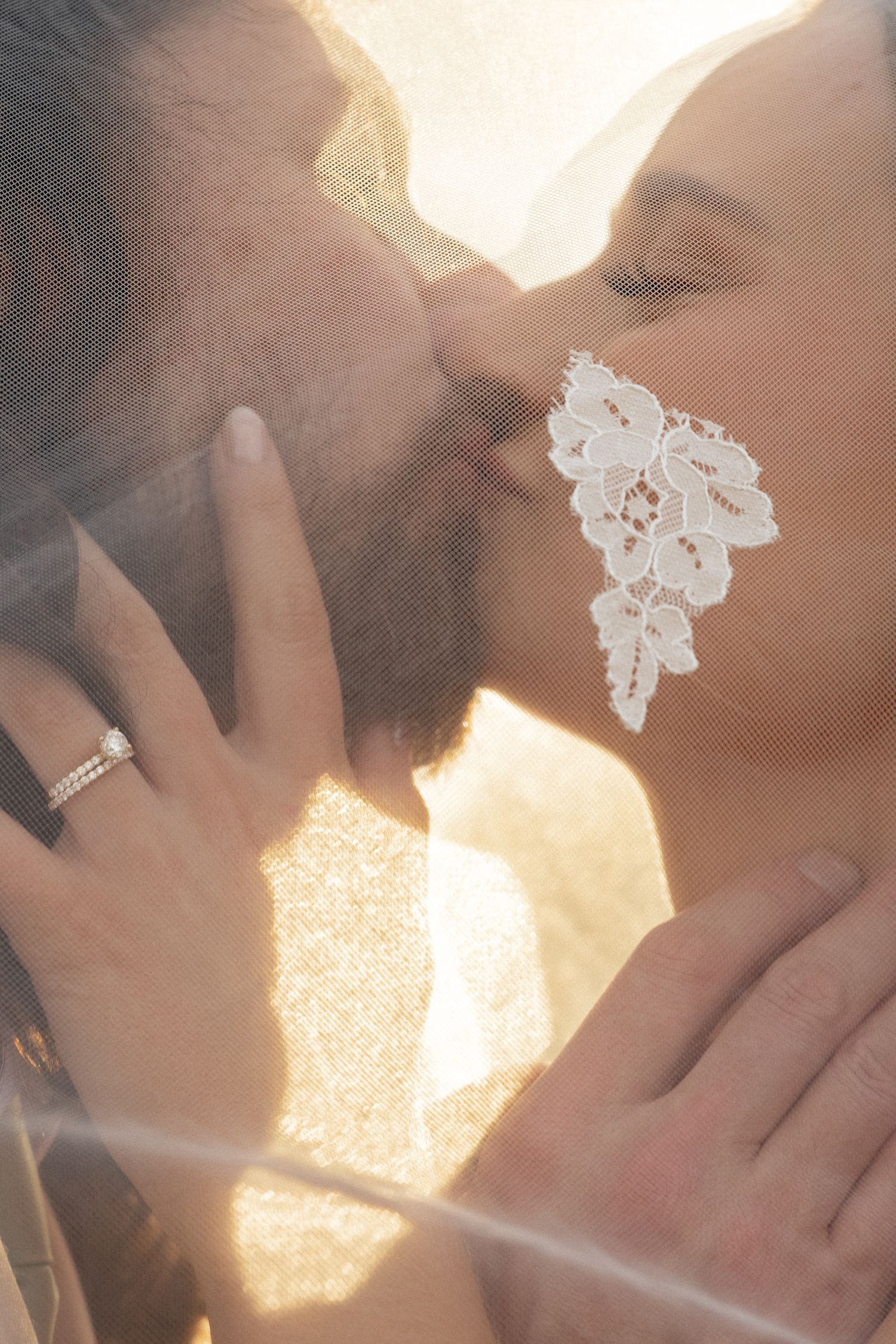 Bride and groom sharing a romantic kiss under a lace veil in golden sunlight, with wedding ring visible.