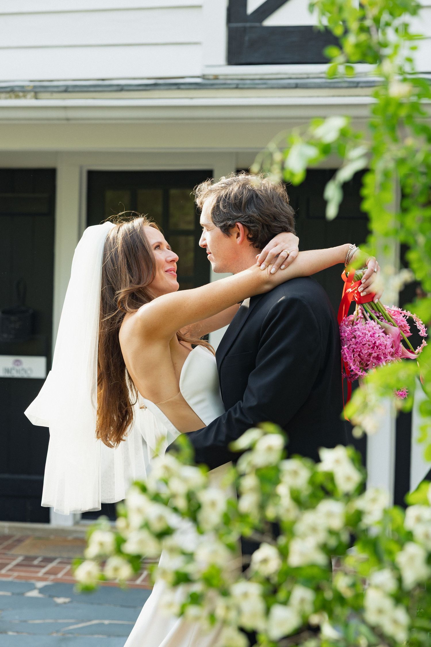 Bride and groom embracing outdoors, holding pink flowers, wearing wedding attire near a white building with greenery.