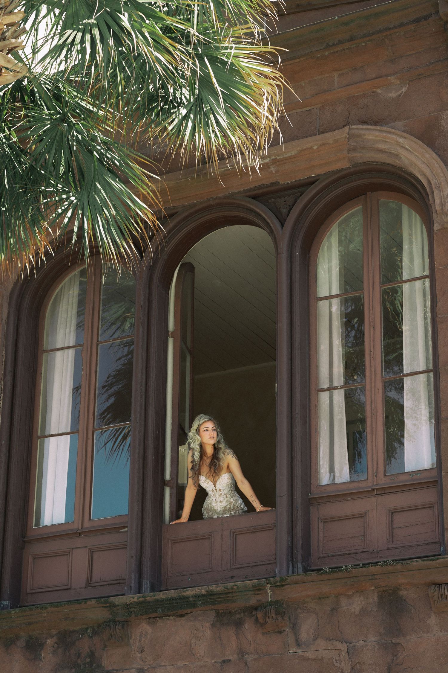 Elegant bride in lace wedding dress gazes from arched wooden window of a historic stone building.
