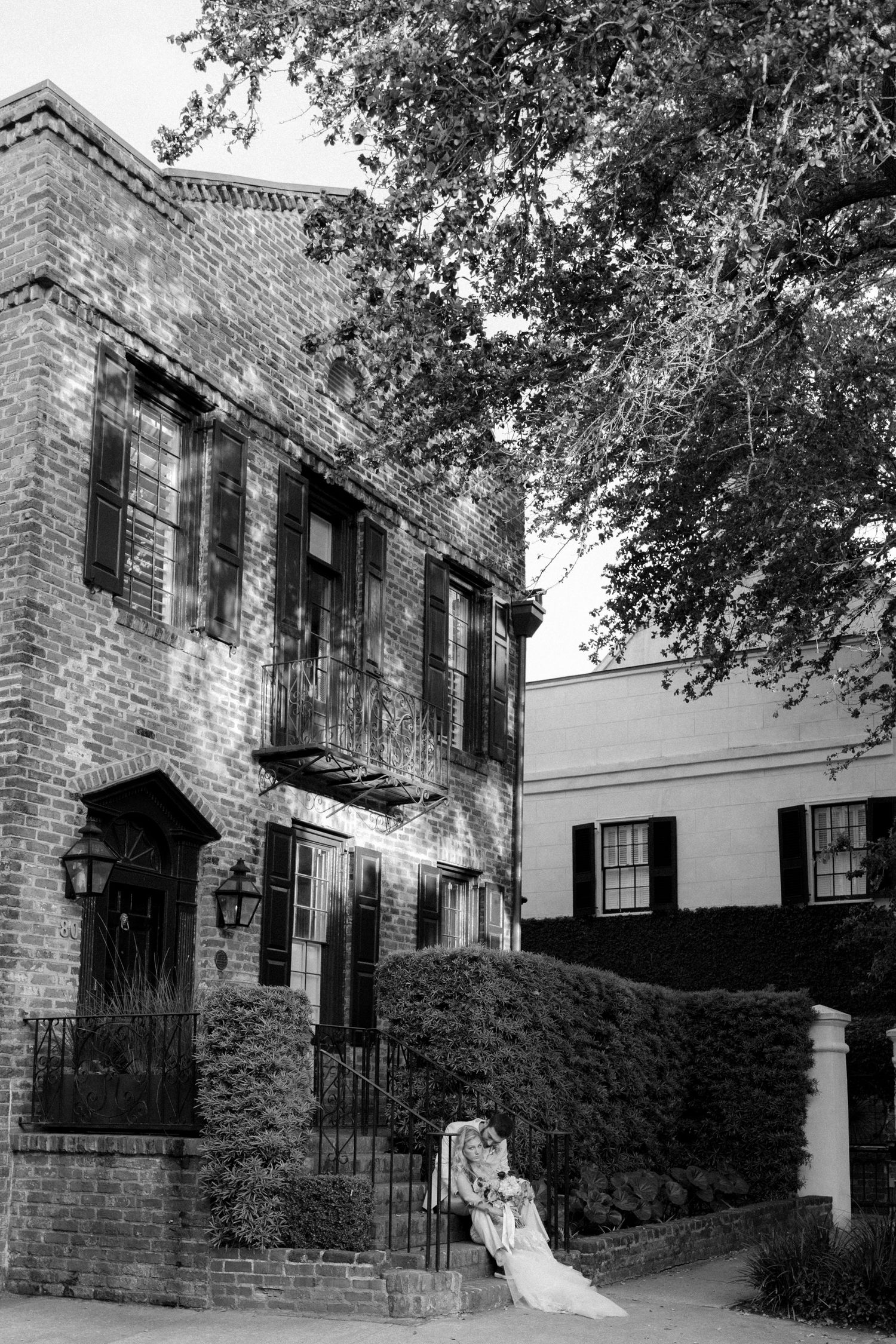 Black and white photo of a bride in a lace gown leaning against a brick building surrounded by lush hedges.