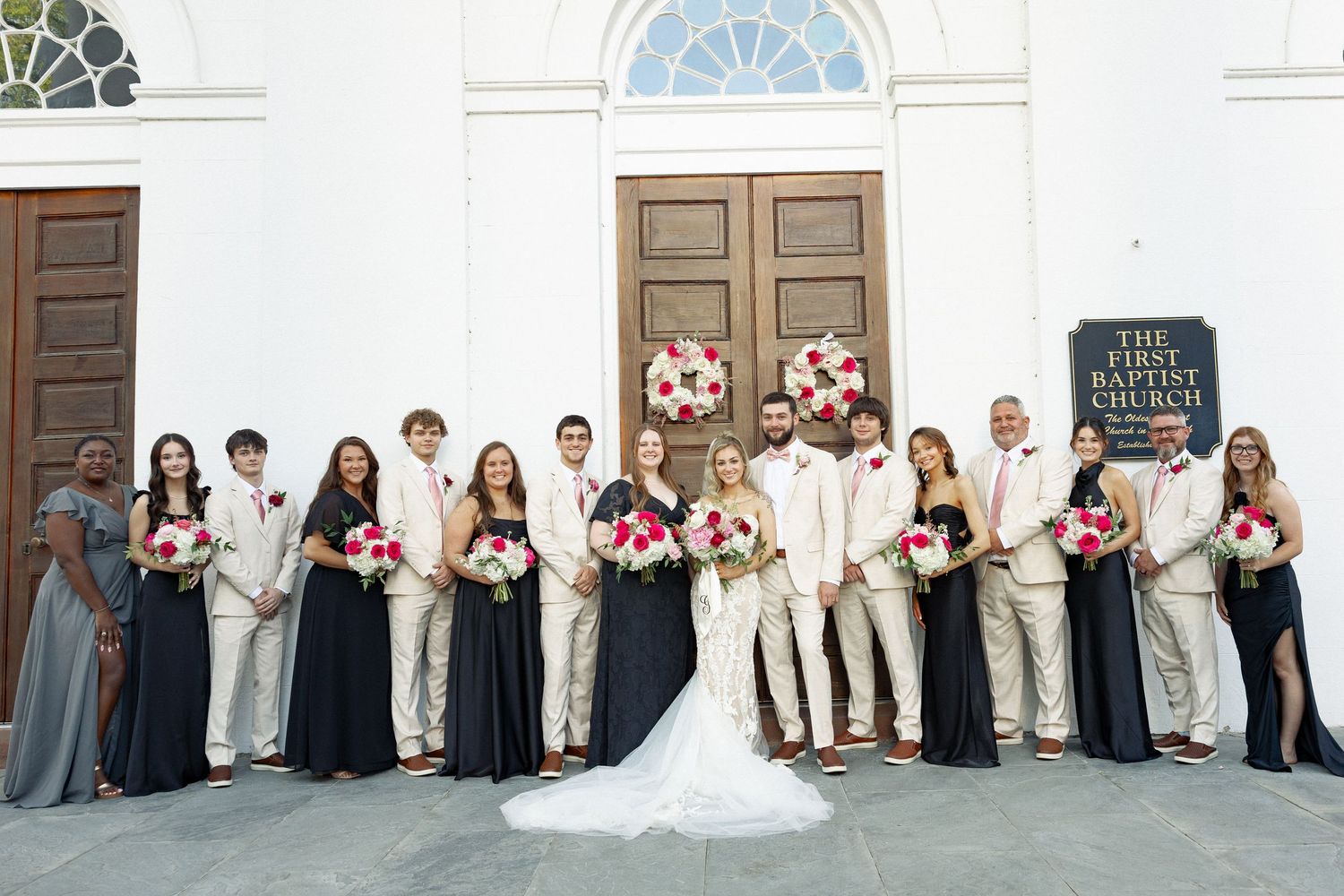Wedding party poses outside First Baptist Church, bridesmaids in navy gowns holding pink bouquets with groomsmen in tan suits.