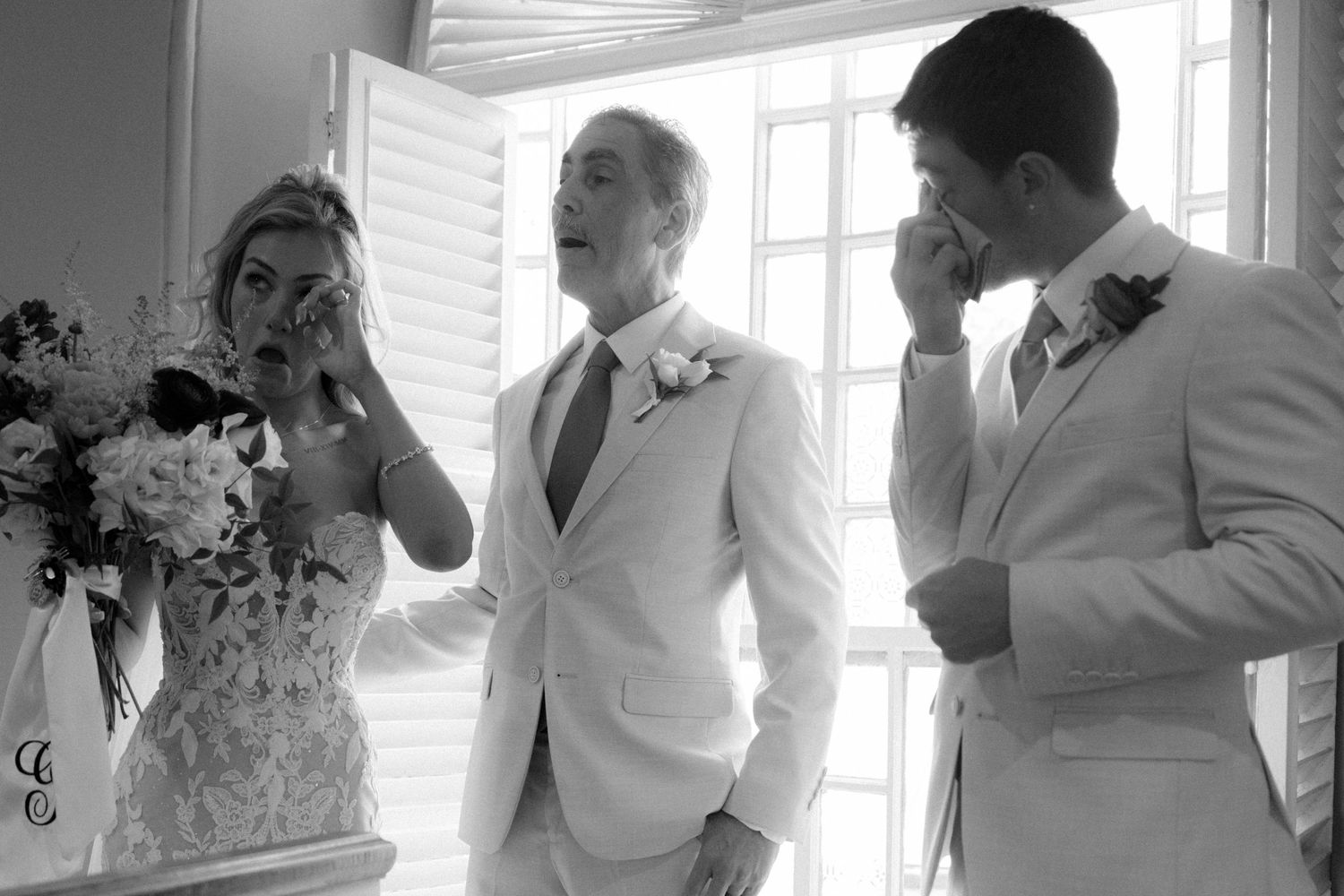 Black and white wedding photo of bride and groom wiping tears with officiant smiling between them.