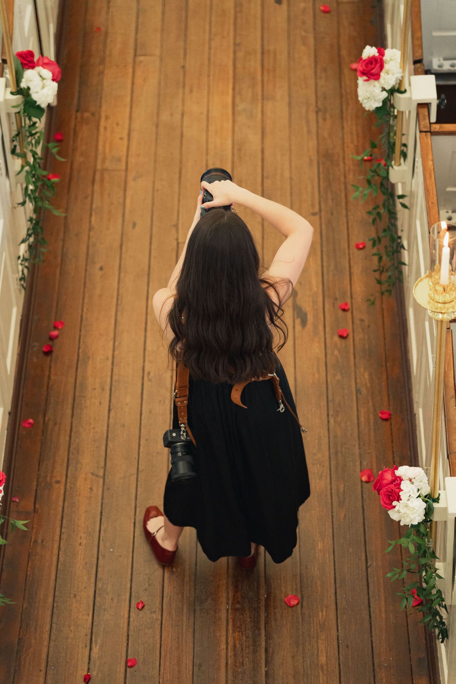 Aerial view of a photographer in black dress capturing the flower-petal-lined wooden church aisle.