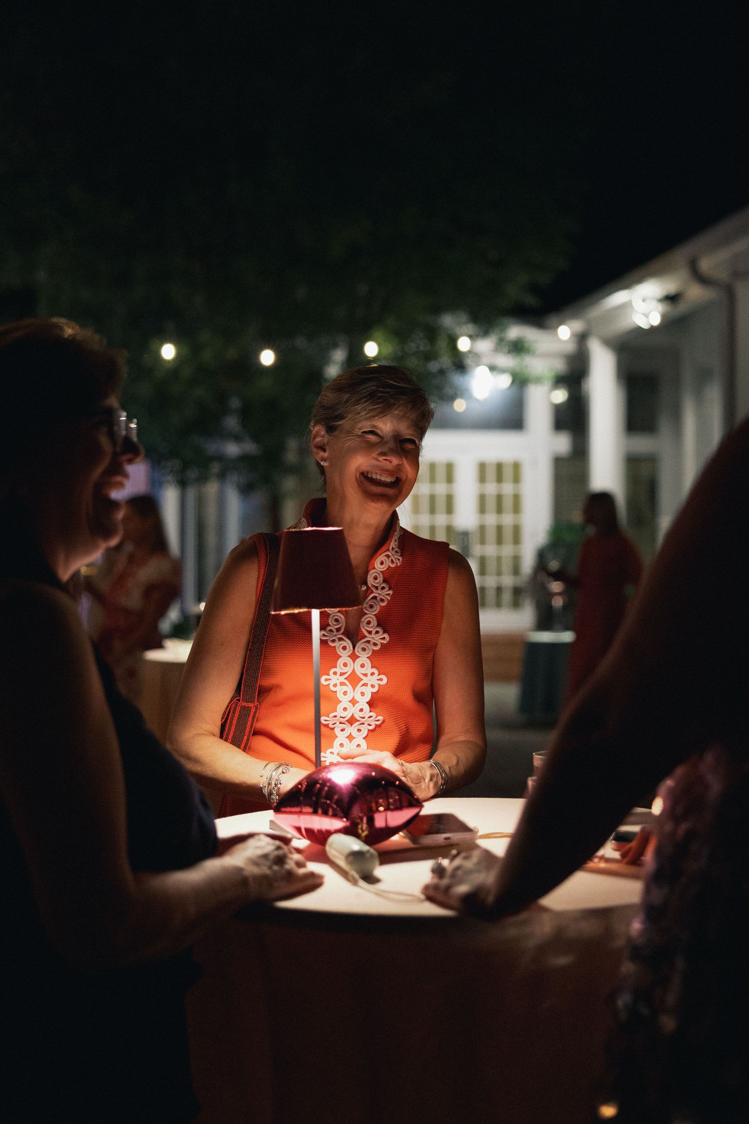 Smiling woman in orange dress at outdoor evening party, illuminated by glowing red centerpiece on table.