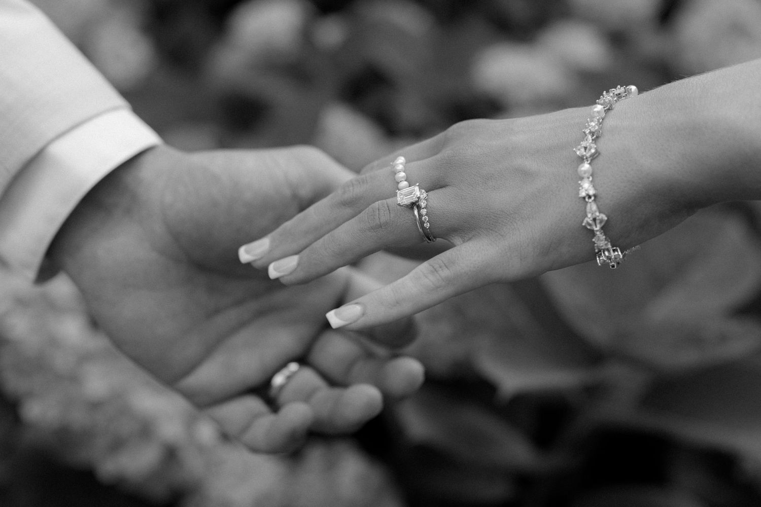 Black and white close-up of bride and grooms hands showcasing diamond engagement ring and tennis bracelet.