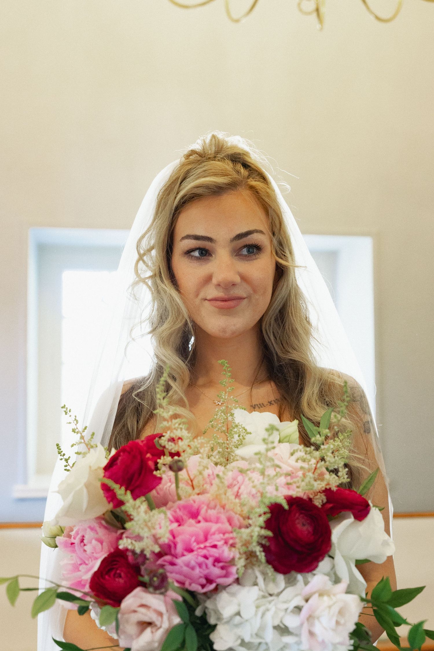 Smiling bride wearing veil holding large bouquet of pink and red peonies and roses indoors.