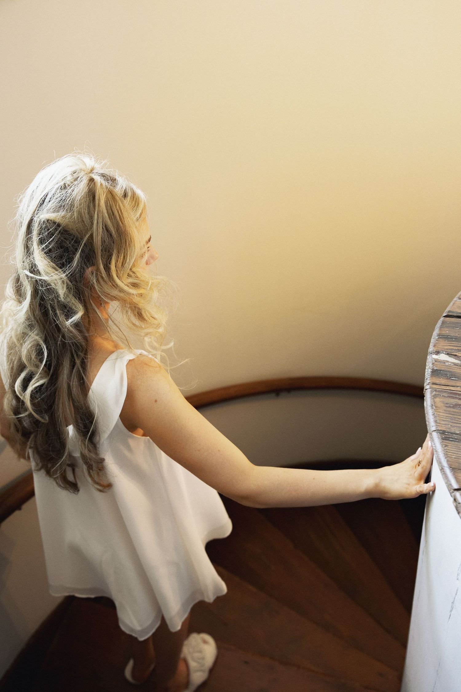 A blonde bride with veil and long curls descends a spiral staircase in a white slip dress.
