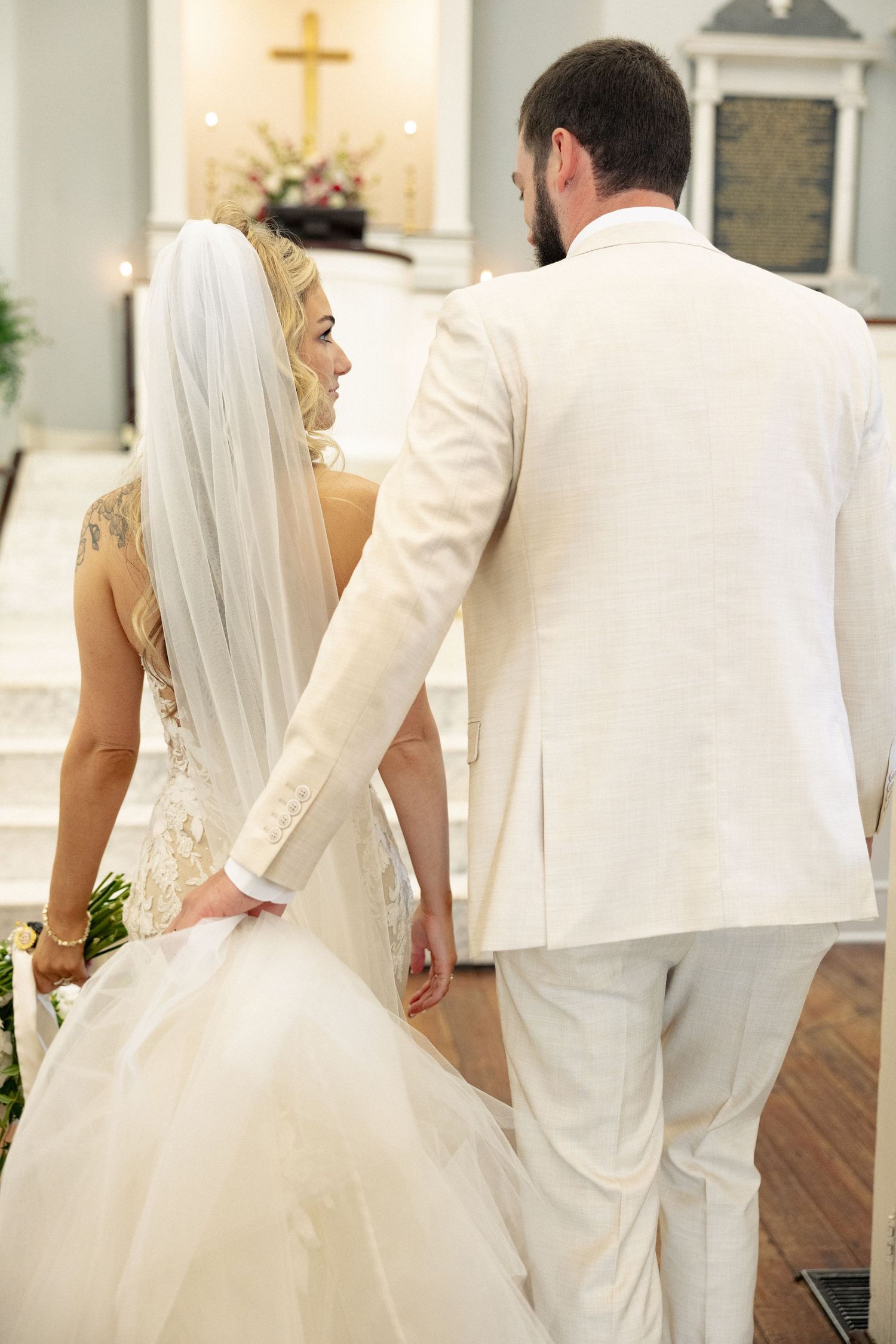 Bride and groom walking toward church altar in white wedding attire, bride wearing veil and holding bouquet.