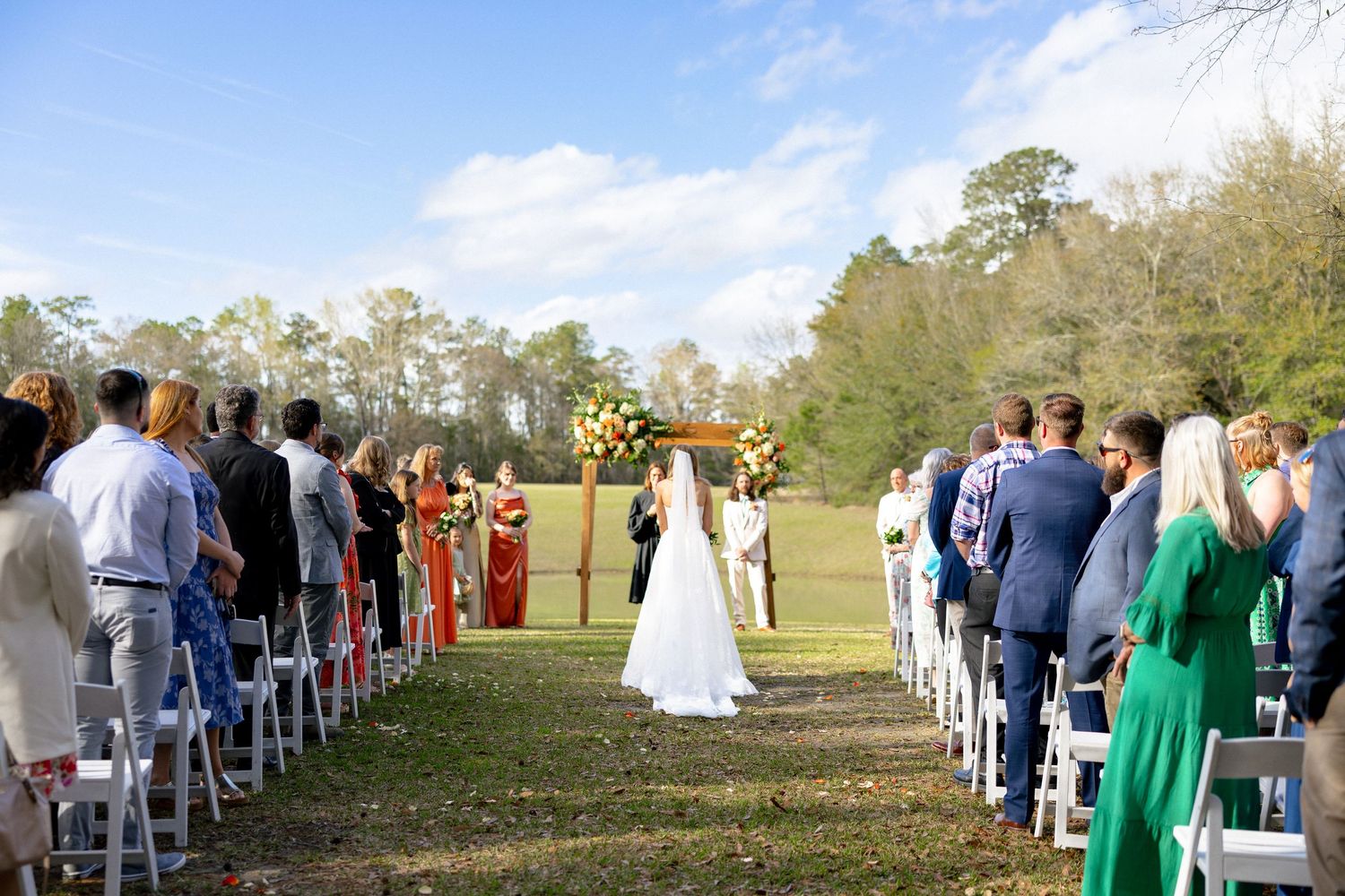 Bride walking down the aisle at outdoor wedding ceremony with guests seated on white chairs in green field.