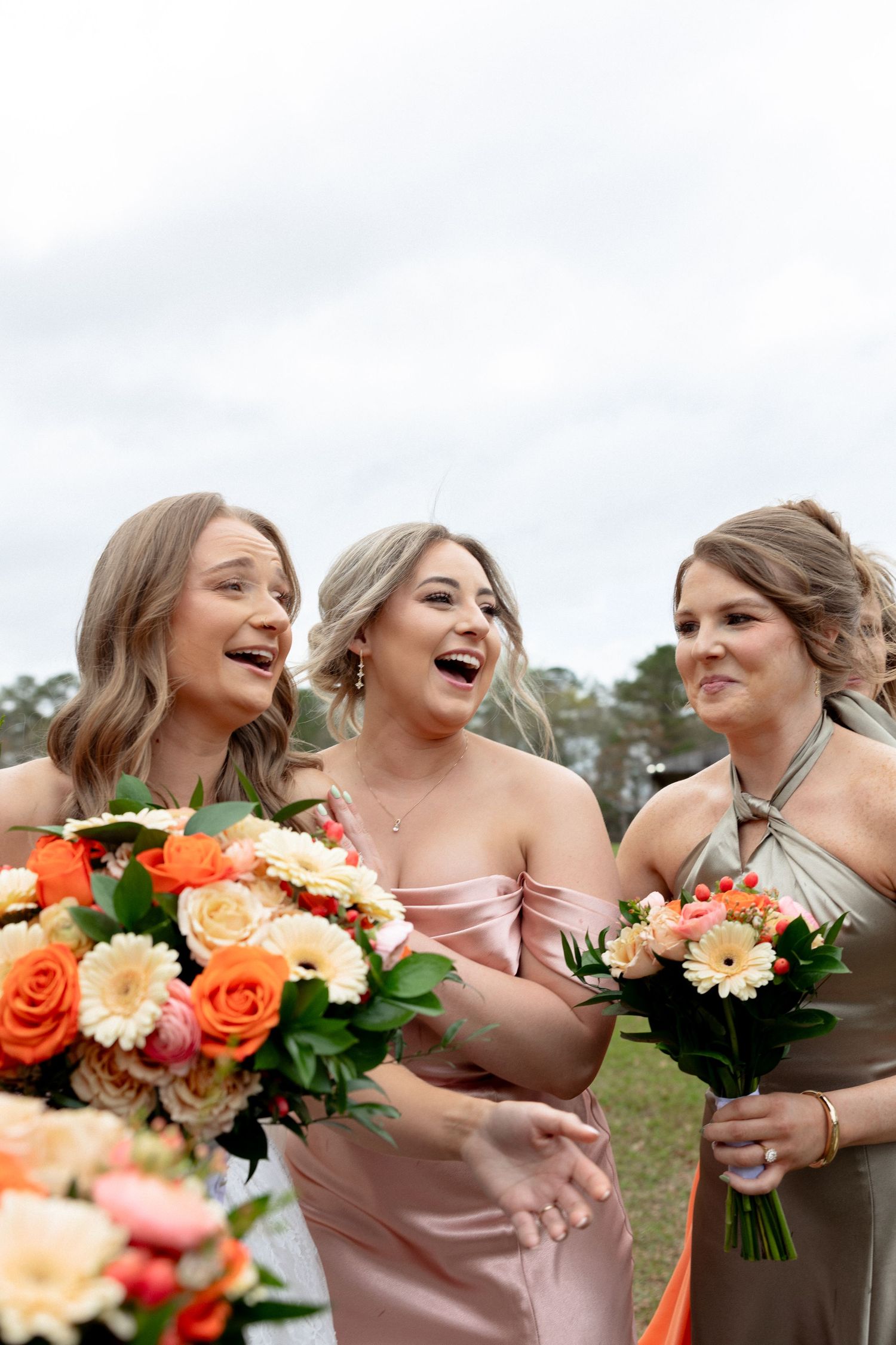 Three bridesmaids in blush and sage satin dresses laugh together while holding vibrant orange and peach bouquets.