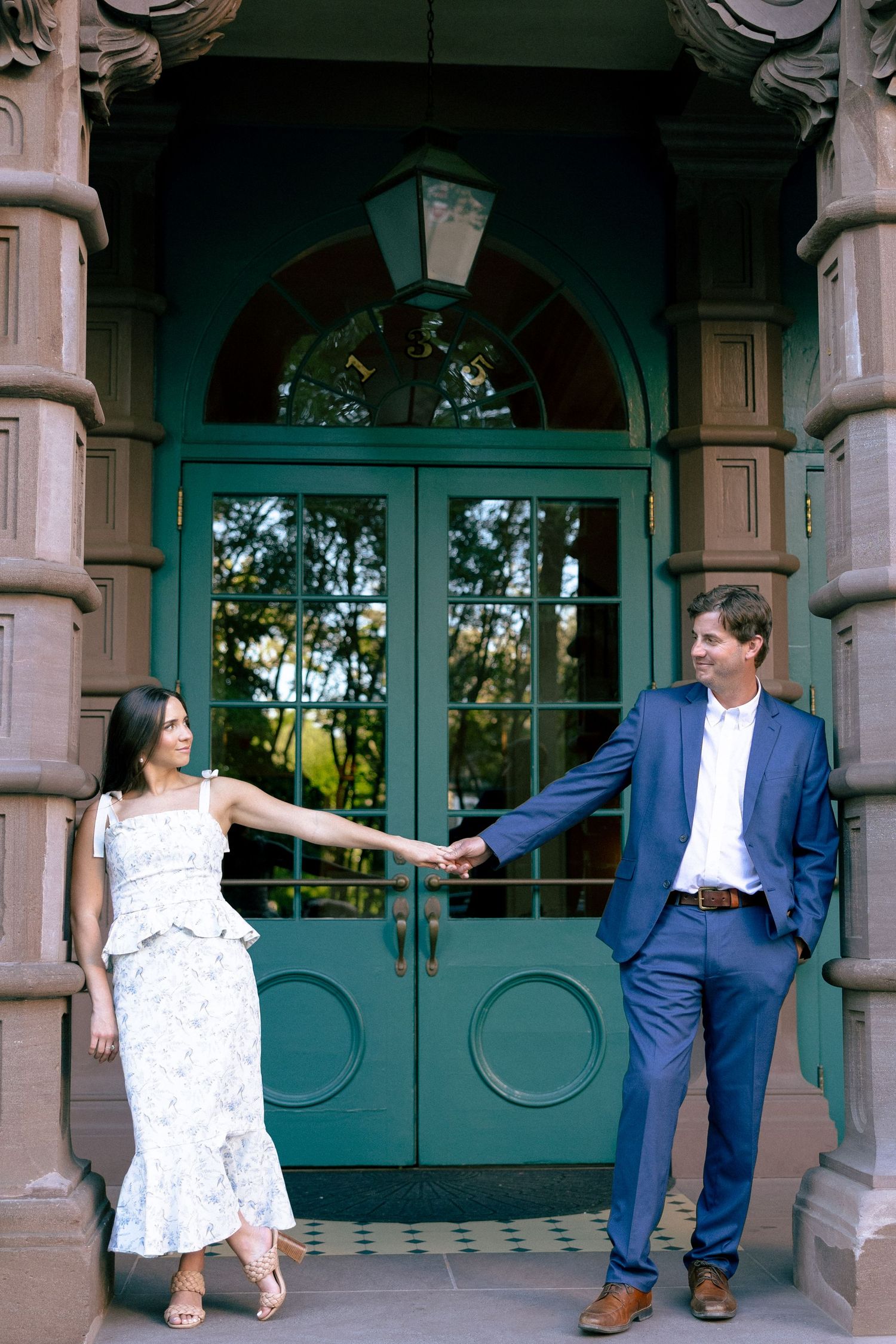 Couple holding hands in front of elegant teal double doors, woman in floral dress, man in navy suit, romantic pose.