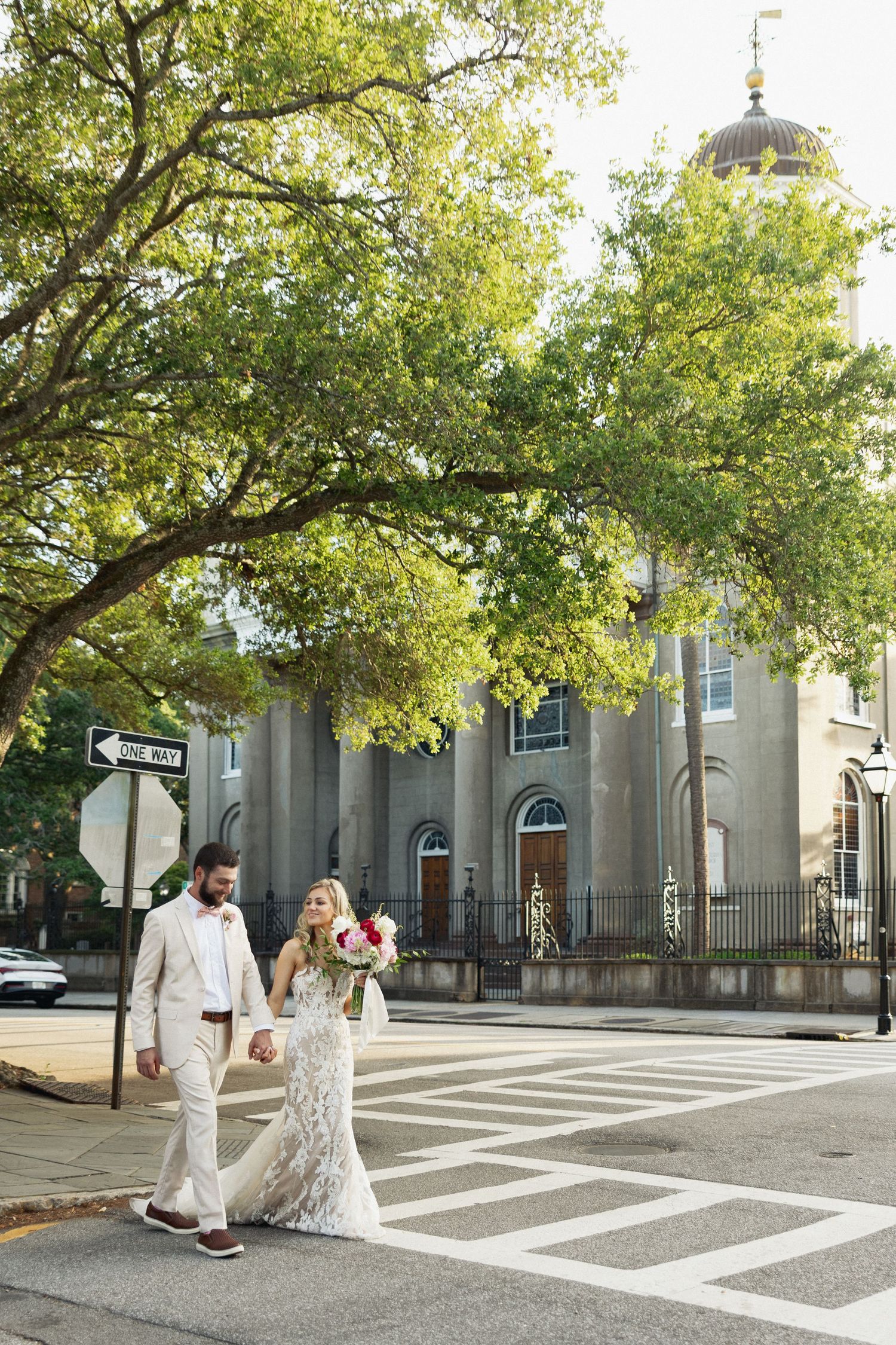 Wedding couple crosses street hand-in-hand near historic domed church building beneath large oak trees in Charleston.