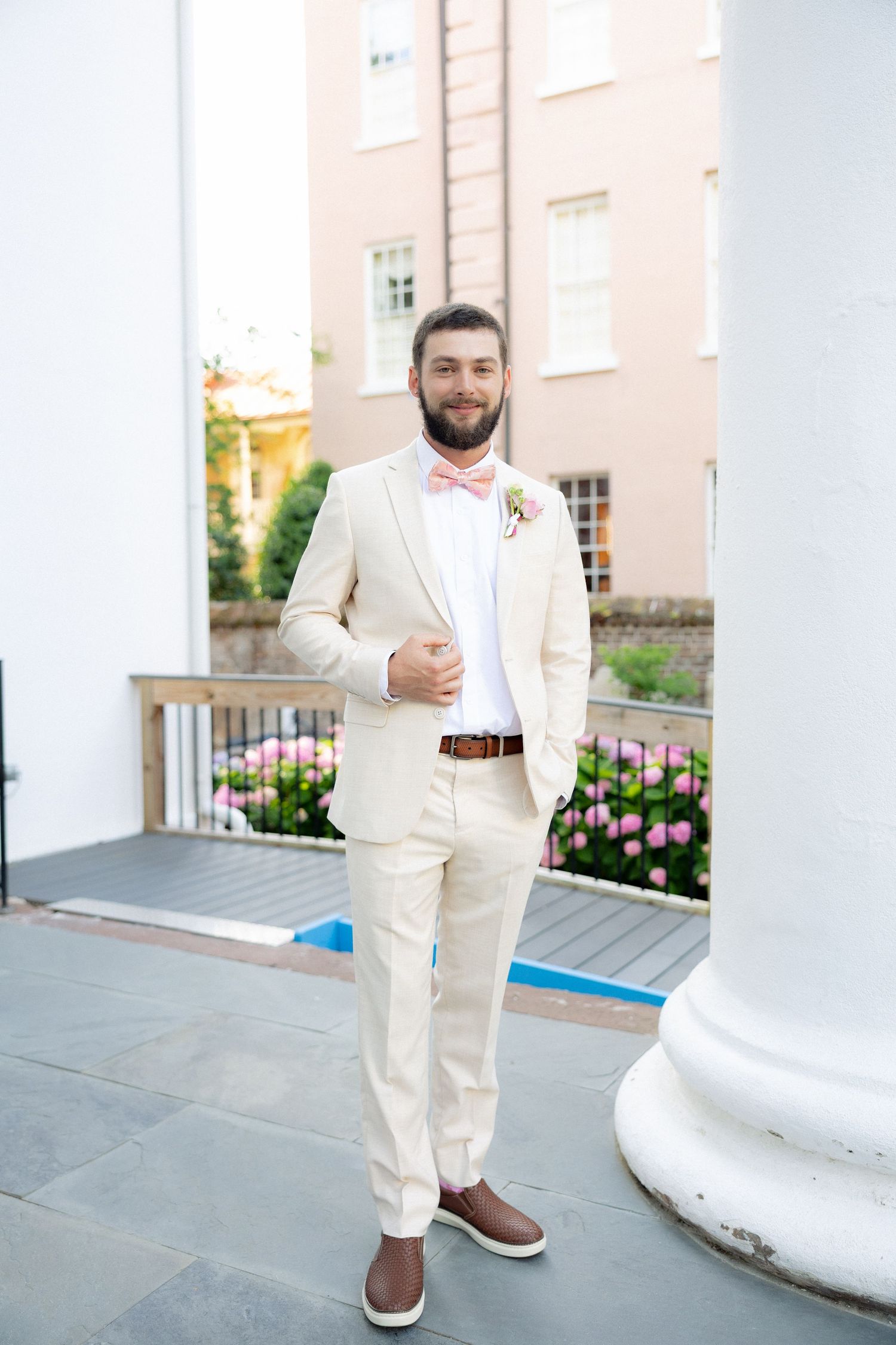Groom in cream suit with pink bow tie and boutonniere stands confidently by white column at outdoor wedding venue.