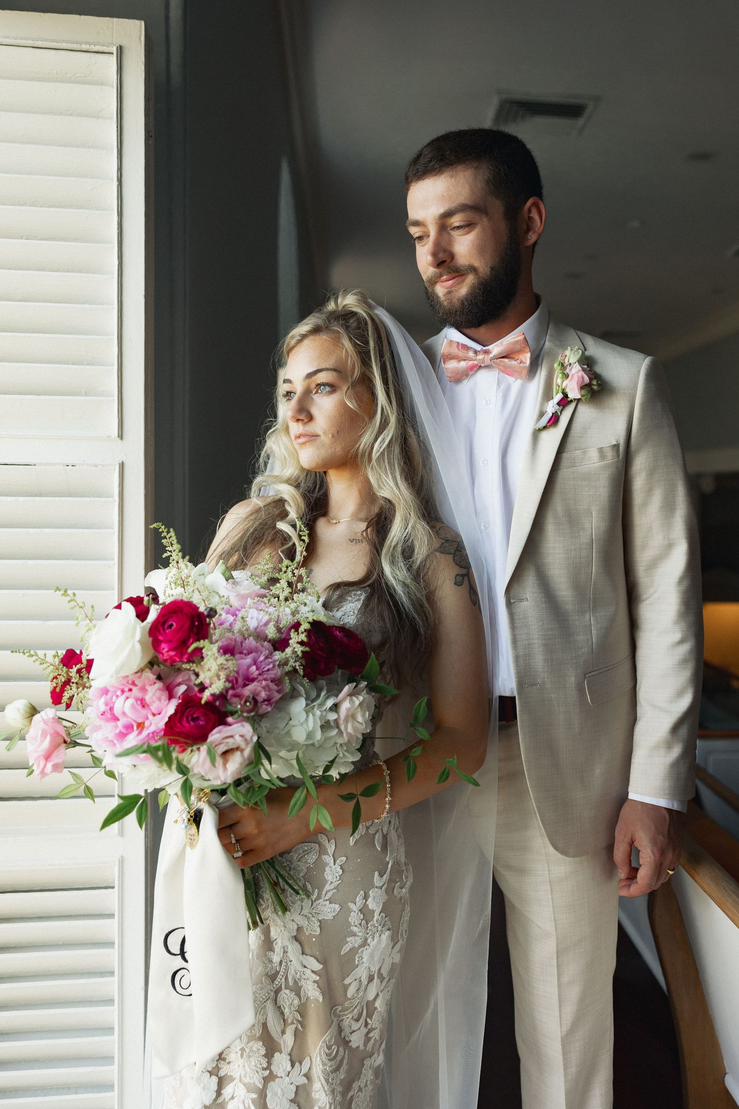 Bride and groom posing indoors, bride holding vibrant pink bouquet, groom in beige suit with pink bow tie.