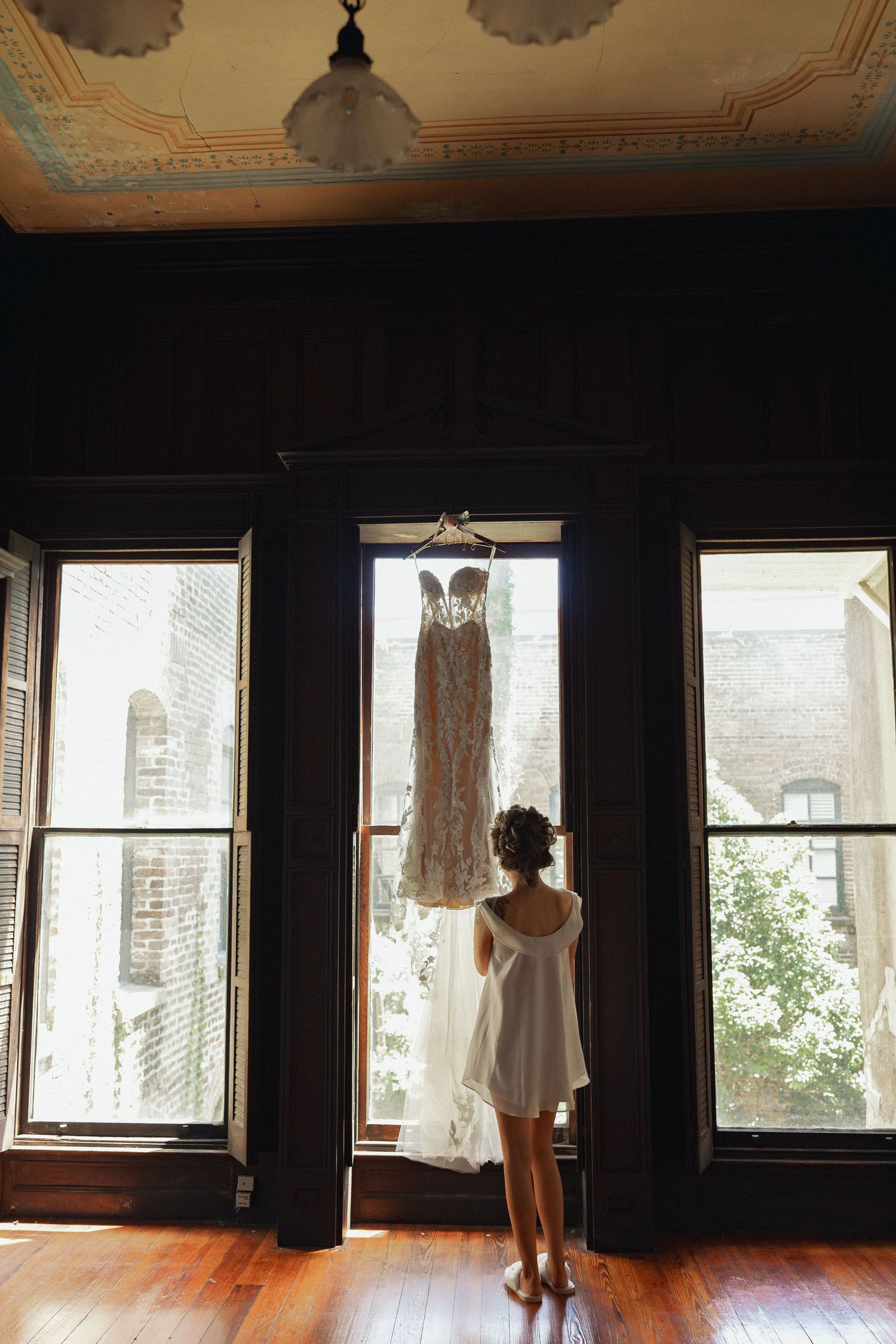 Bride in white slip dress admiring lace wedding gown hanging in sunlit window.