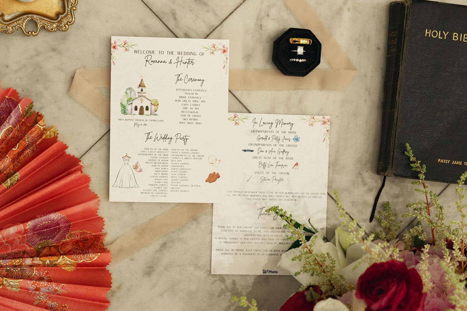 Wedding flat lay with illustrated ceremony programs, red fan, Holy Bible, ring box, and floral accents on marble.