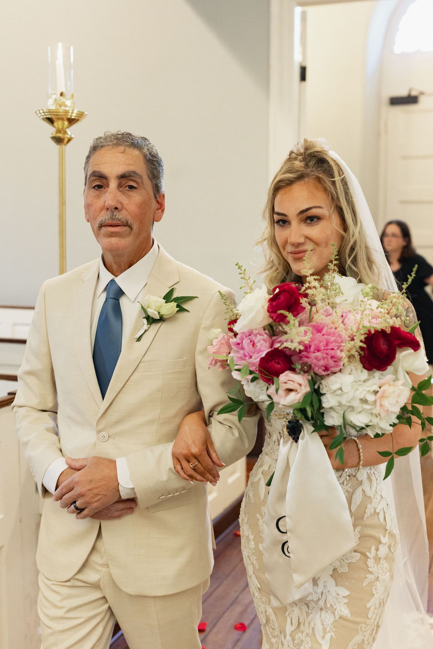 A bride holding a colorful bouquet walks down the aisle with her father in a beige suit and blue tie at a church wedding.