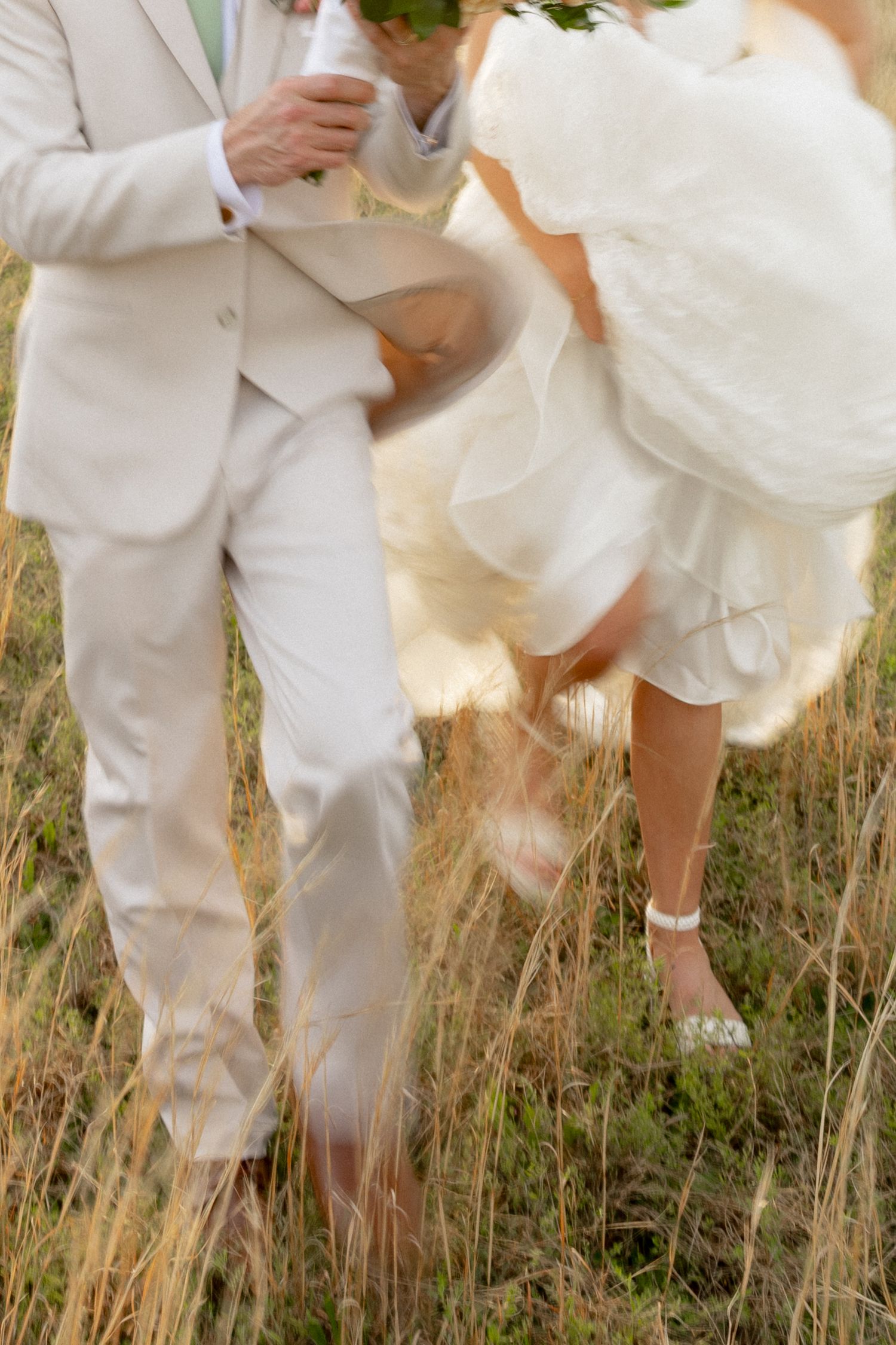 Motion-blur photo of bride and groom playfully running through tall grass field, bride in white lace gown.