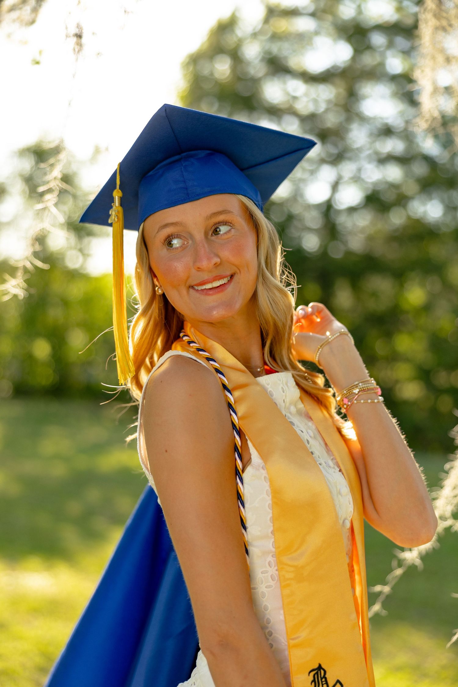 Smiling female graduate wearing blue cap and gold honor cords in sunny outdoor setting.