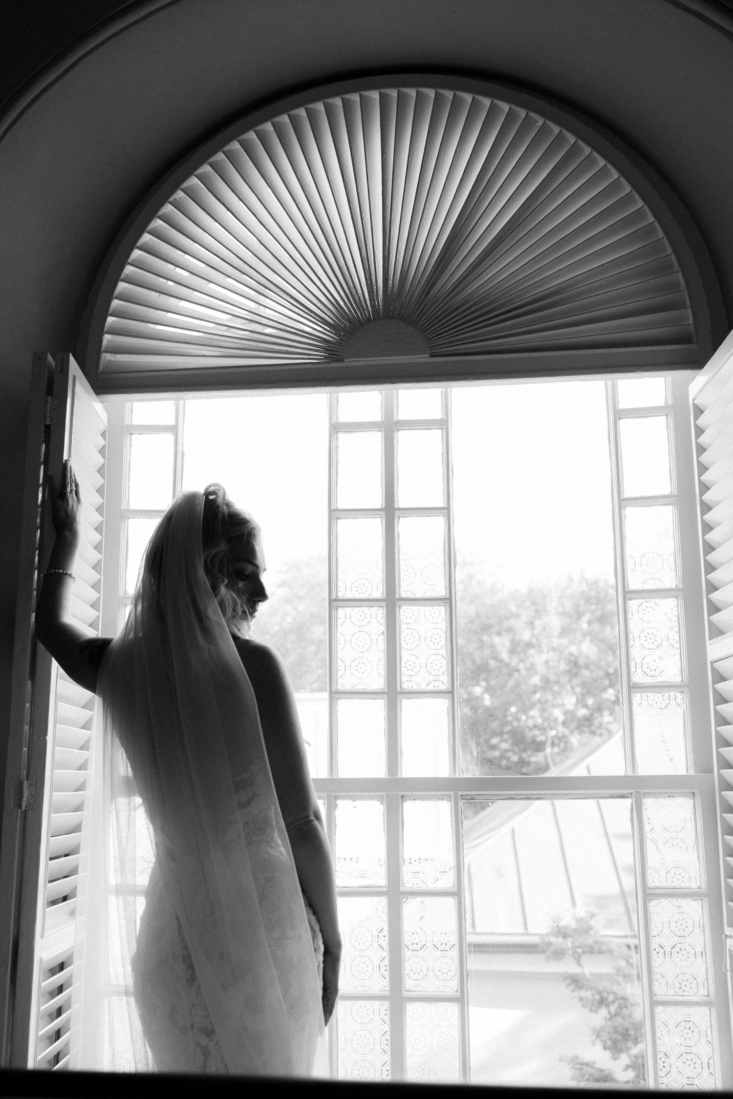 Black and white silhouette of bride in veil standing at arched window with decorative fan transom.