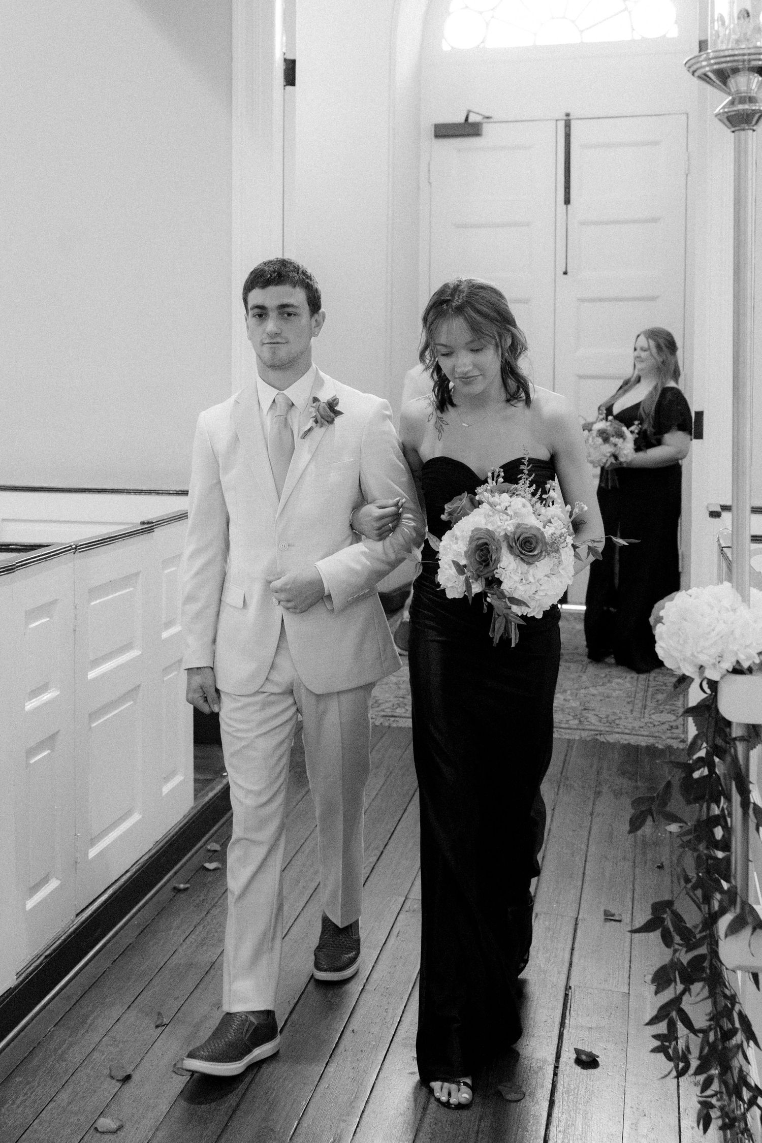 A groomsman in a light suit escorts a bridesmaid in a black gown down the aisle in a church ceremony.