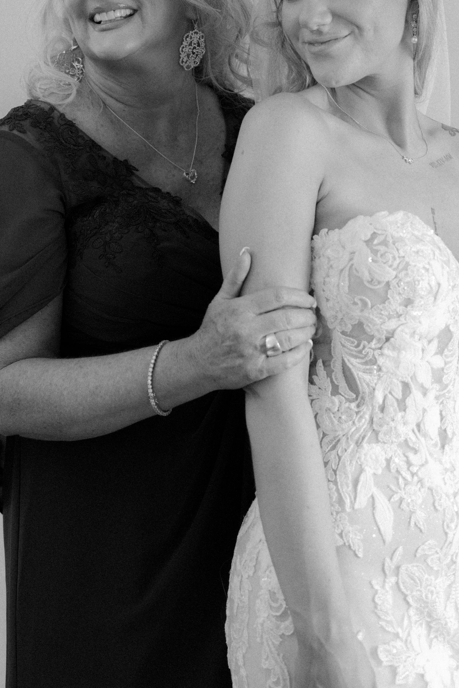 A black and white photo of a mother in a black lace dress embracing a bride in a white gown.