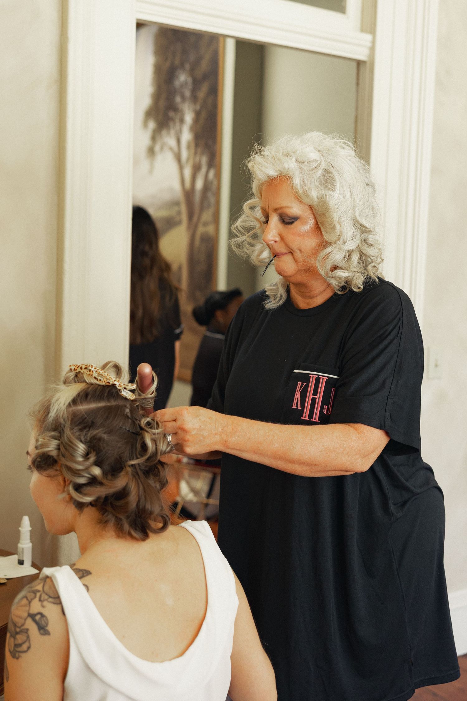 A silver-haired hairstylist in a black monogram shirt styling a seated woman's curly updo before a mirror.