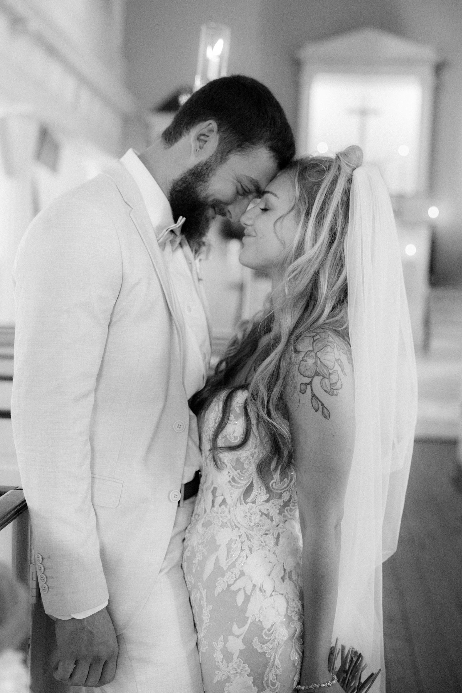 Black and white photo of a bride and groom sharing an intimate moment in a church, foreheads touching.