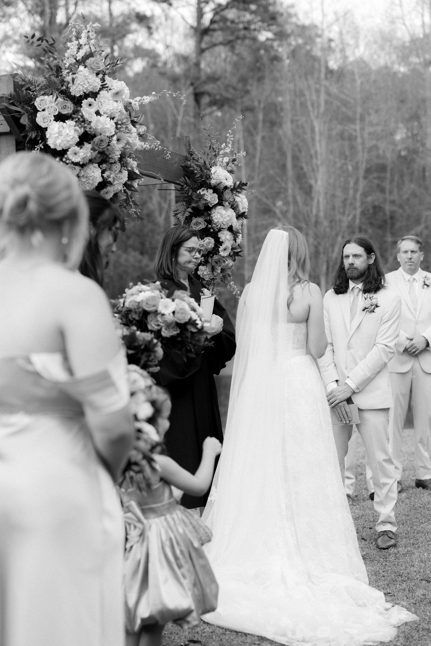 Black and white photo of bride approaching groom at outdoor wedding ceremony beneath floral arch.