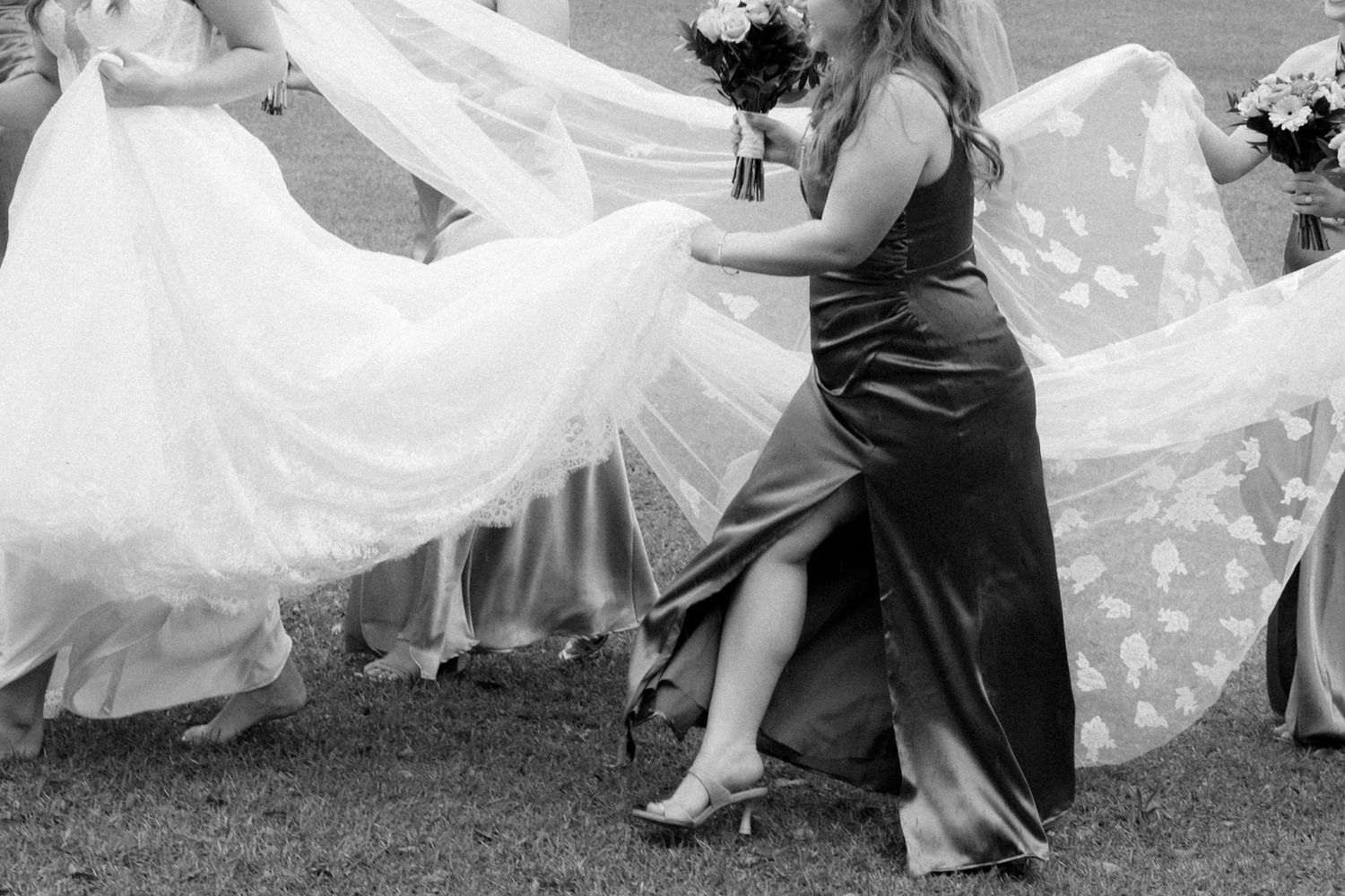 Bridesmaid in dark gown helps carry bride's flowing white lace wedding dress train outdoors in black and white photo.