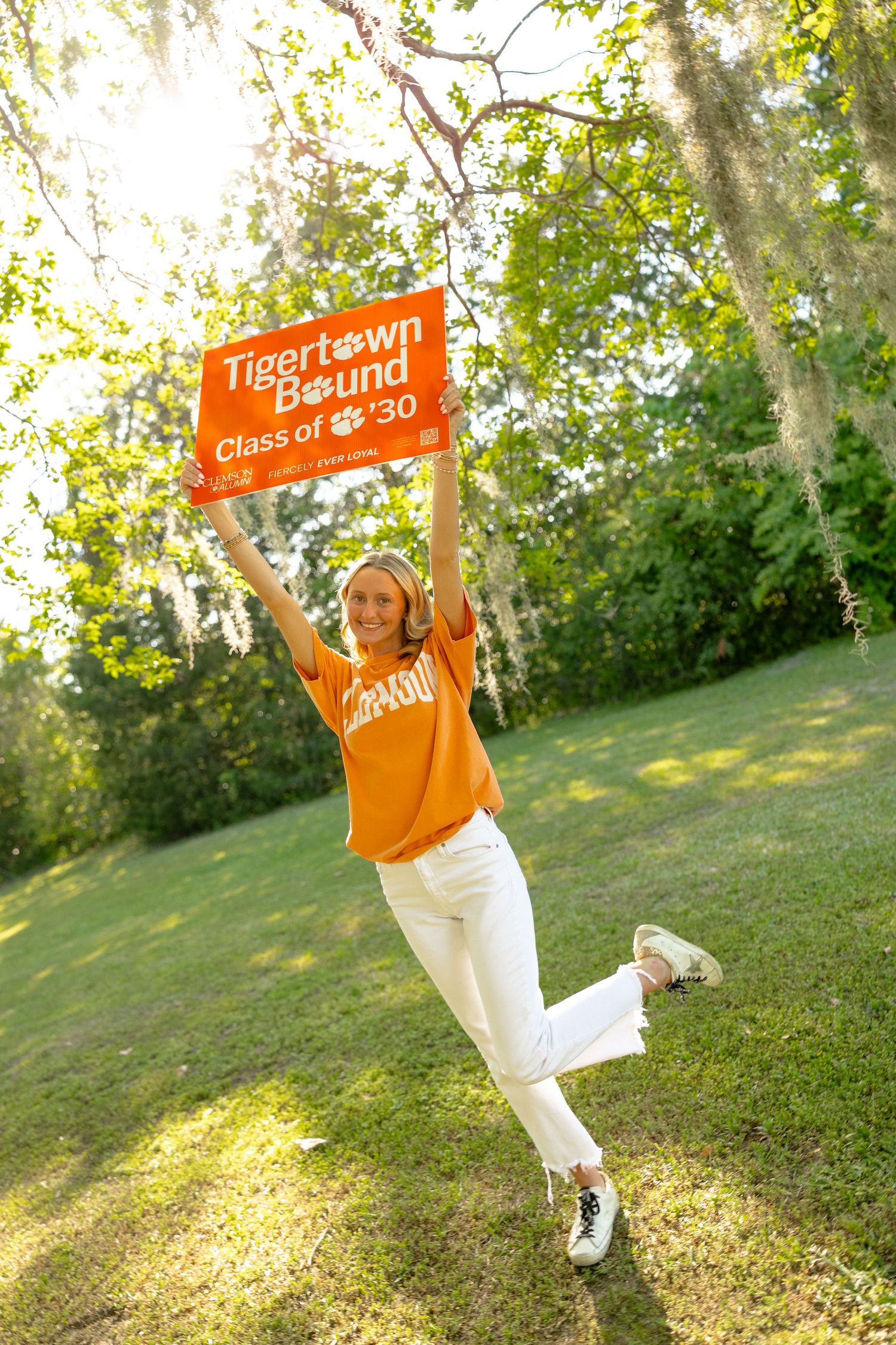 Excited young woman jumping while holding a Clemson Tigertown Bound Class of 2030 orange sign outdoors.
