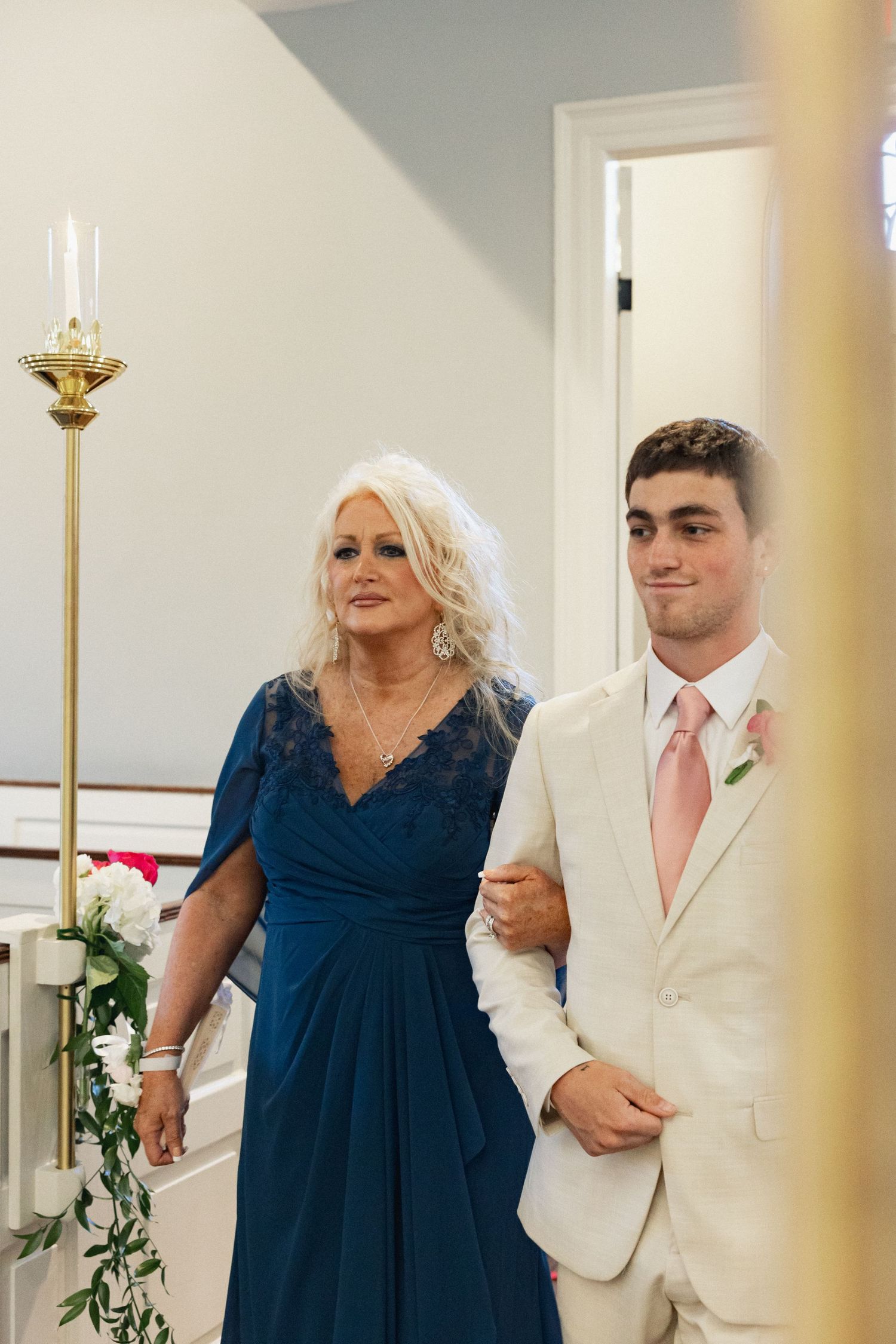 A woman in a blue lace dress walks down the church aisle escorted by a young man in a beige suit with a pink tie.