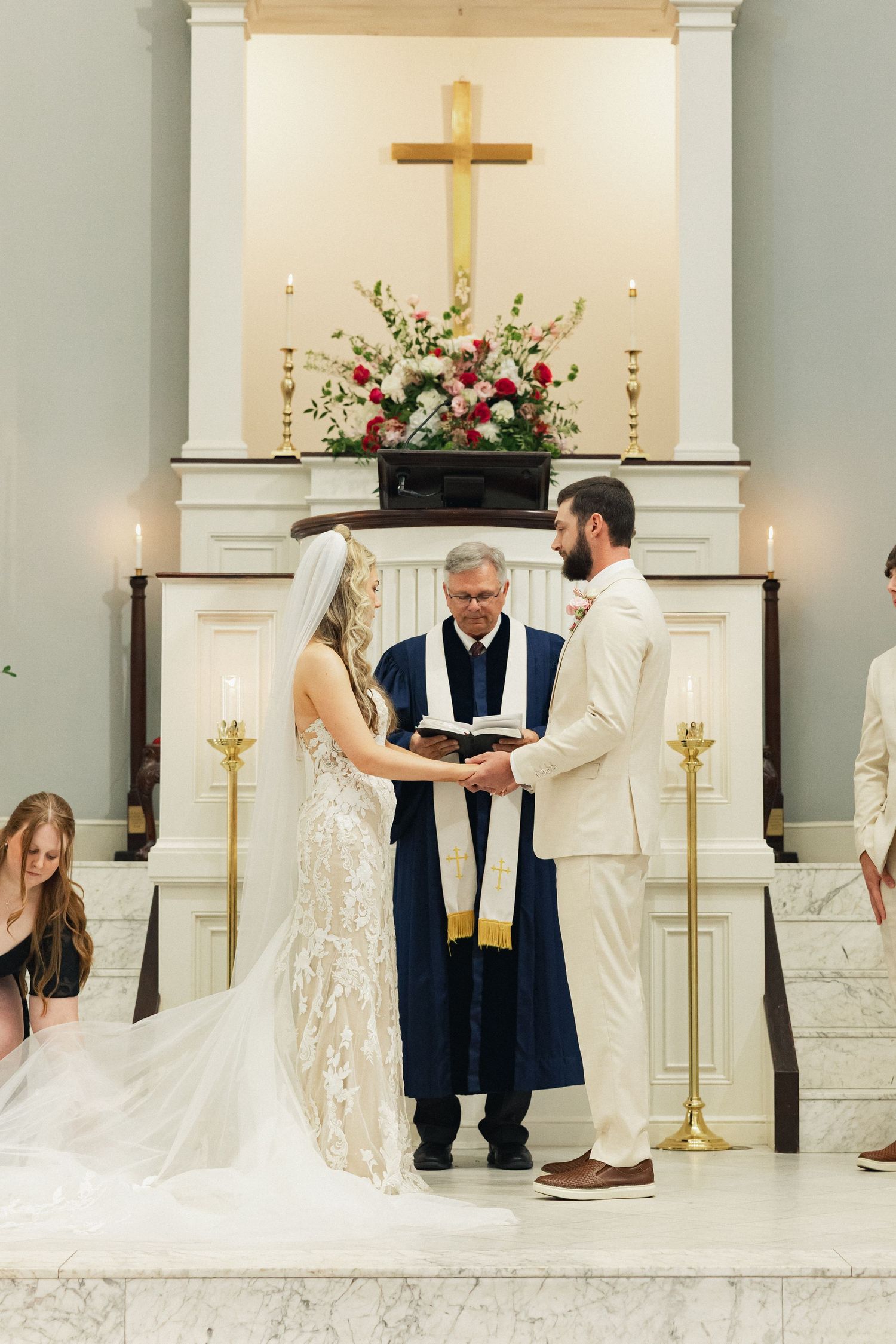 Bride and groom exchange vows before officiant at church altar adorned with cross and floral arrangement.