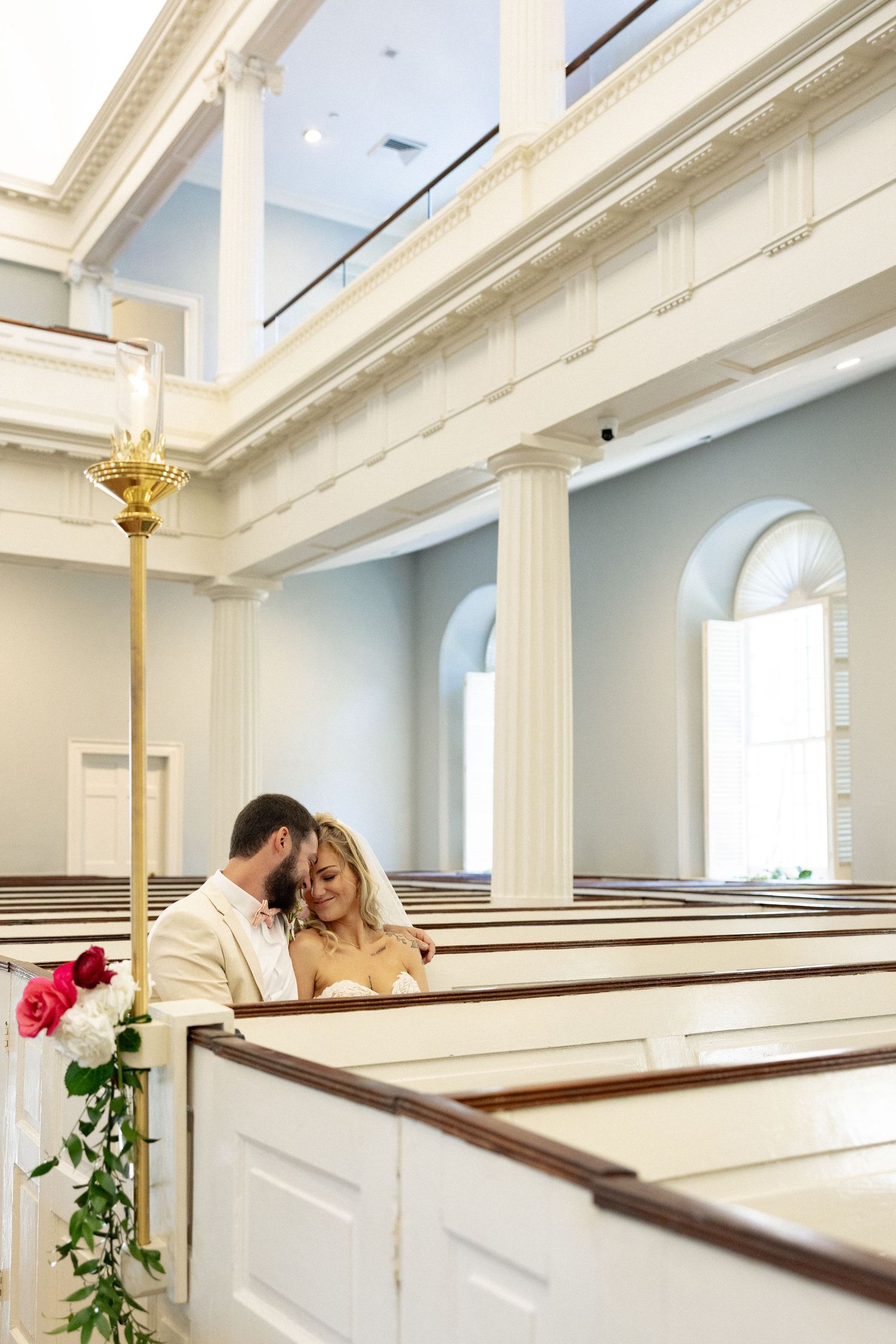 Bride and groom share a romantic moment in a elegant white church with tall columns and wooden pews.