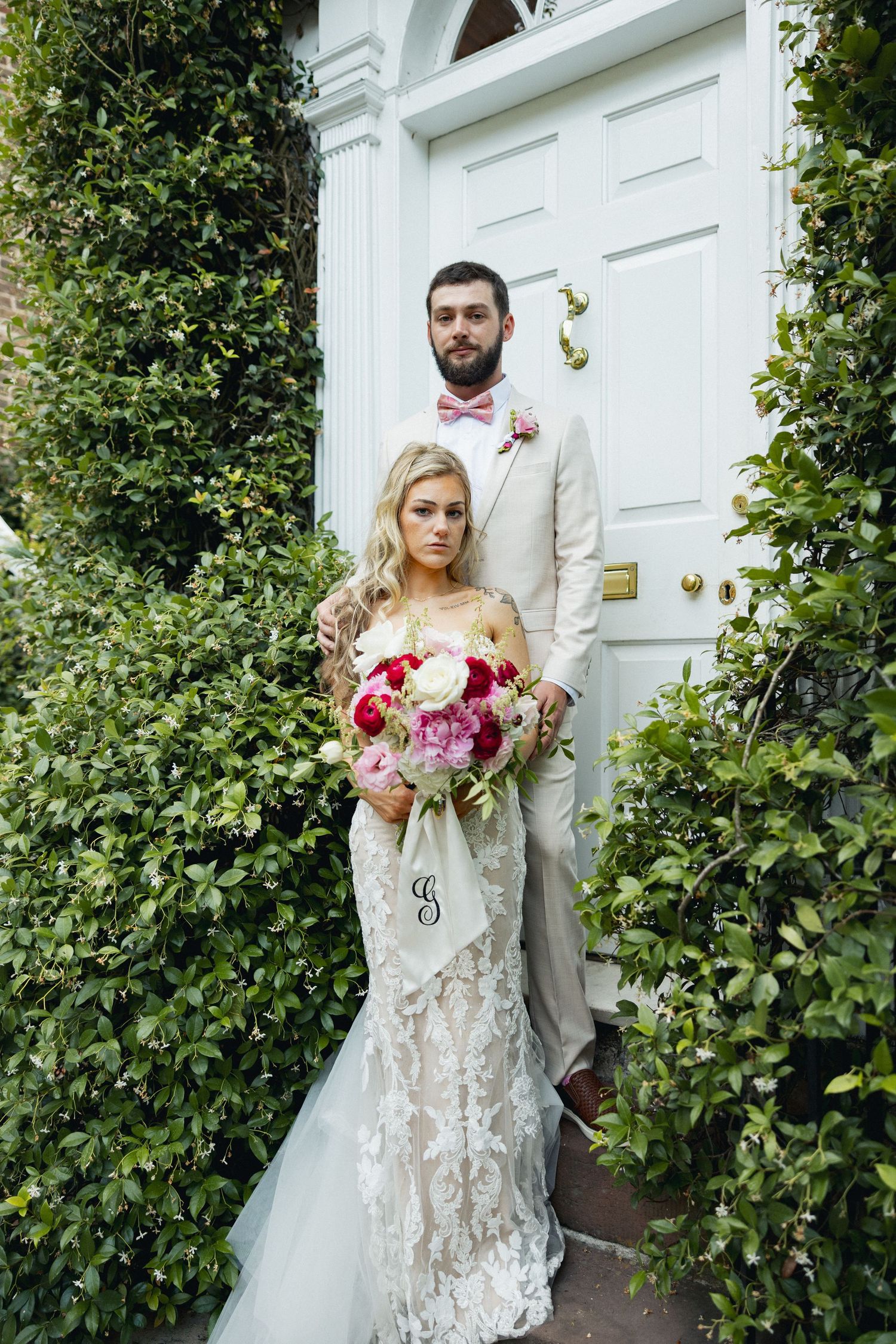 Bride and groom standing before white door framed by greenery, lace gown, tan suit, pink bow tie, monogrammed bouquet.