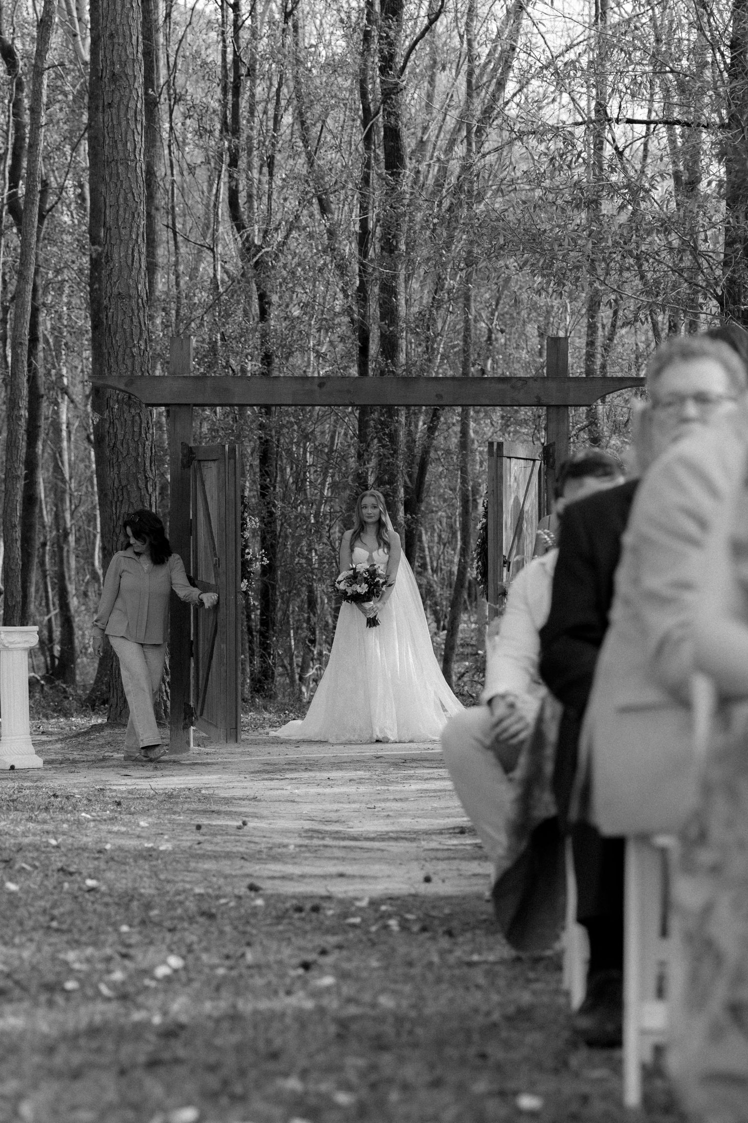 Black and white photo of a bride in white gown approaching the altar through a wooden arch at an outdoor forest wedding.