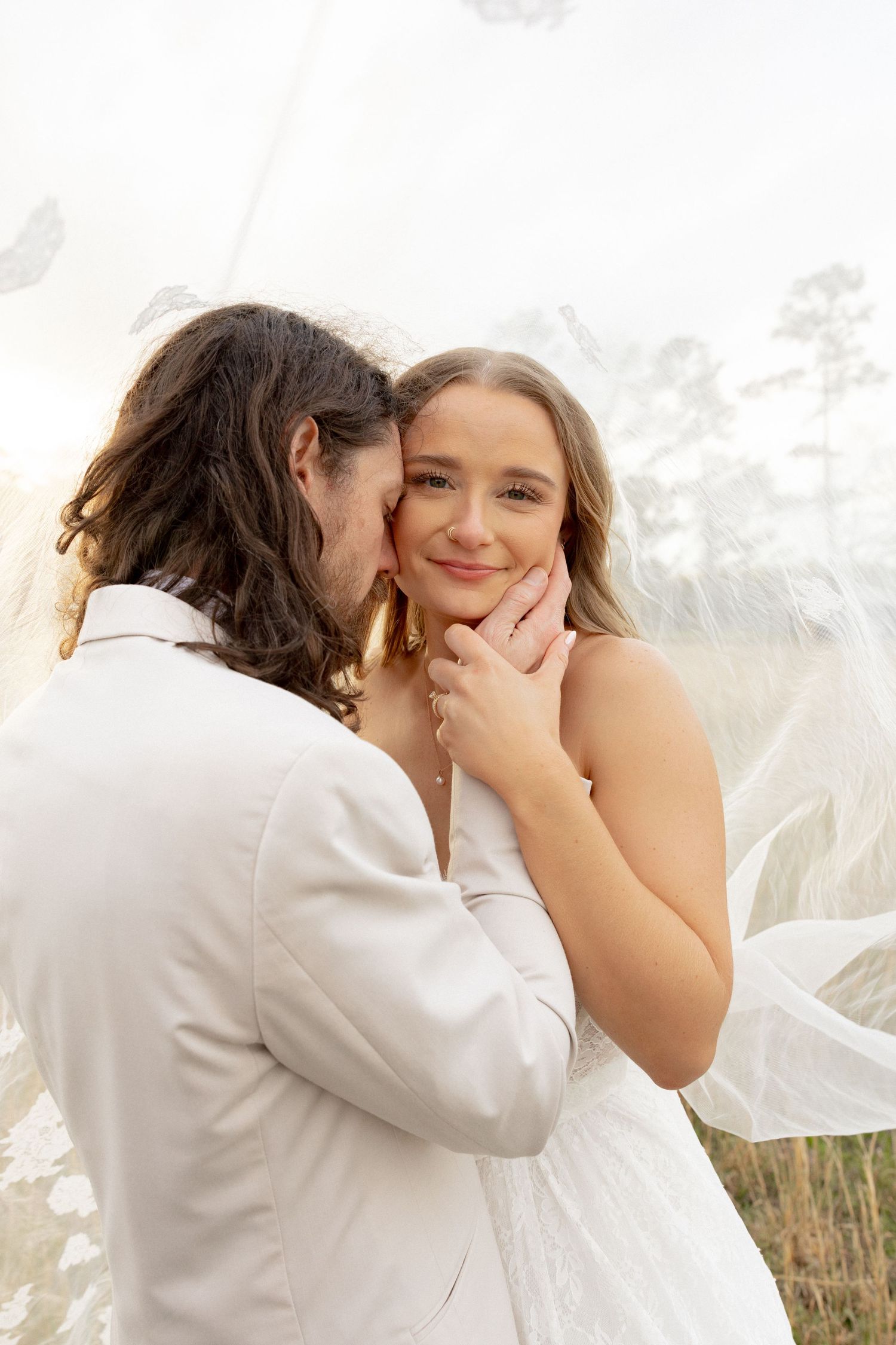 Groom tenderly kisses bride's cheek as she smiles under flowing lace wedding veil.