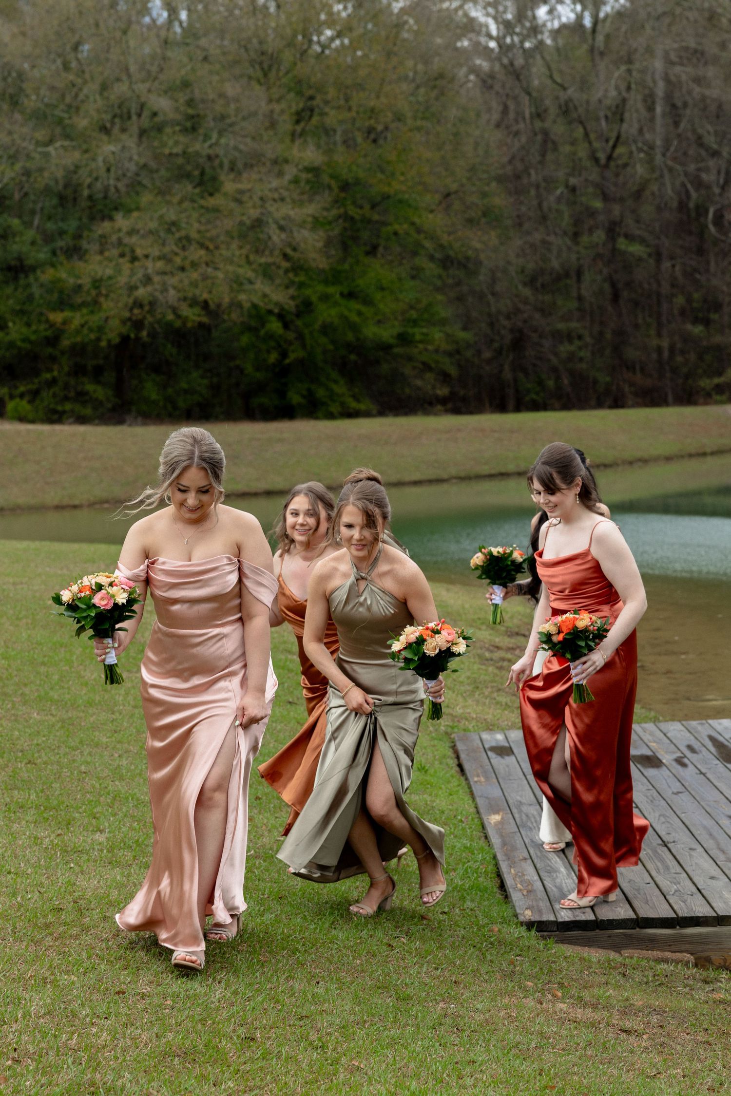 Four bridesmaids in satin gowns of blush, olive, and rust laugh while walking off a wooden dock onto green grass.