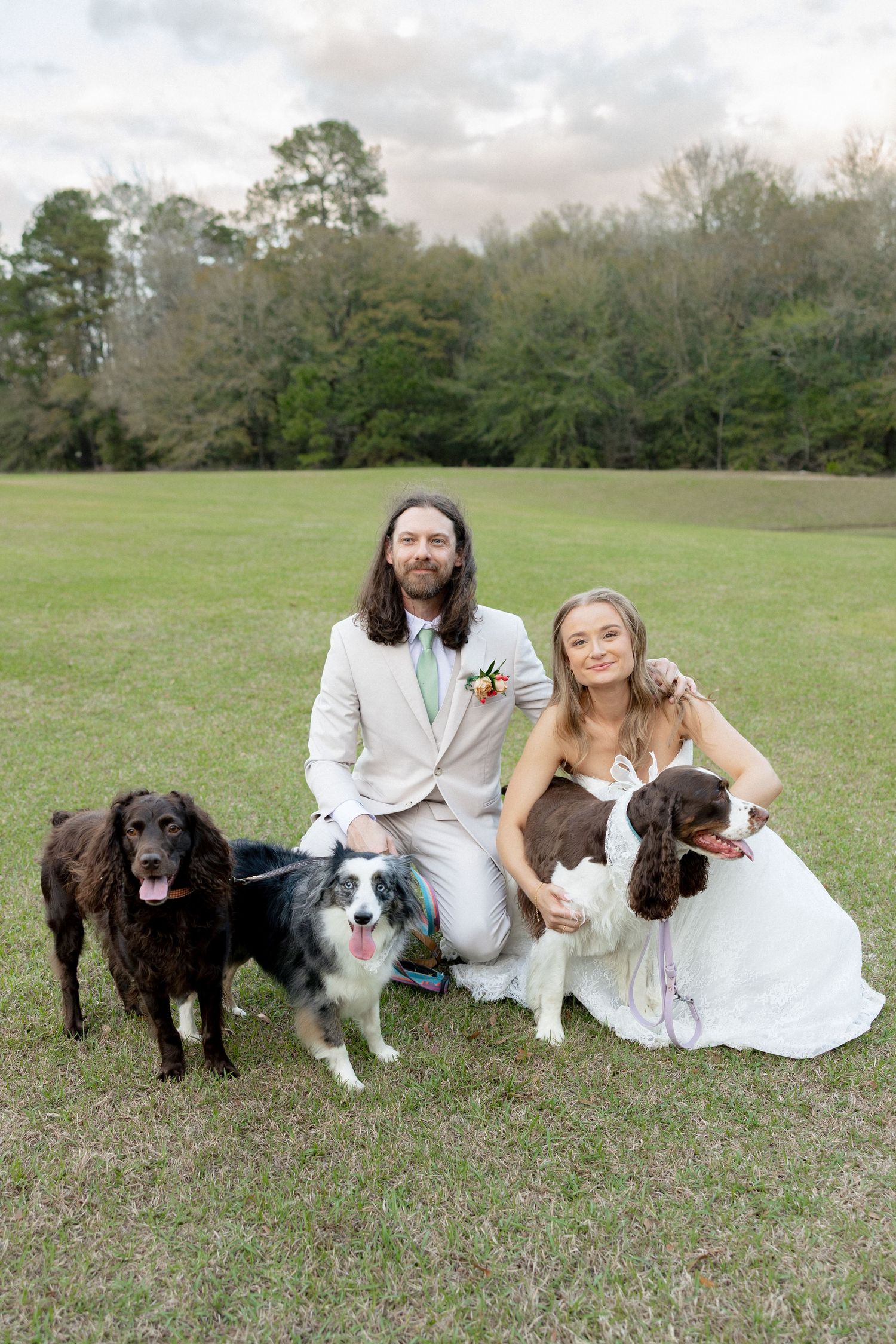 Smiling newlyweds kneeling with three dogs on grass at an elegant outdoor wedding.