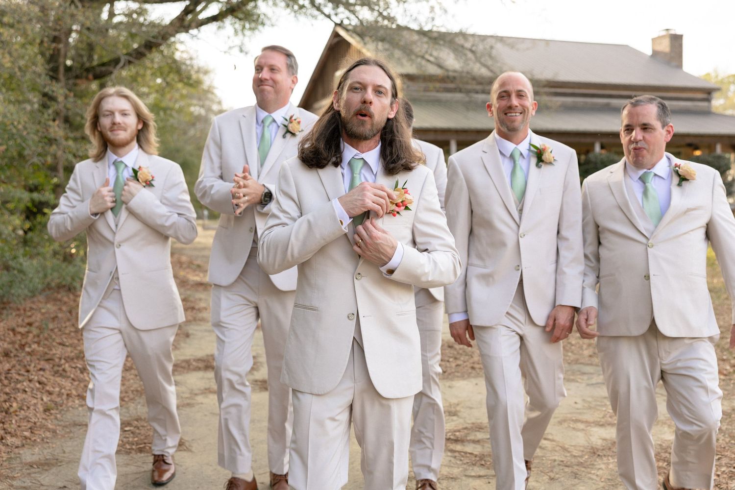 Groom with long hair leads groomsmen in cream suits walking toward camera at outdoor barn wedding venue.